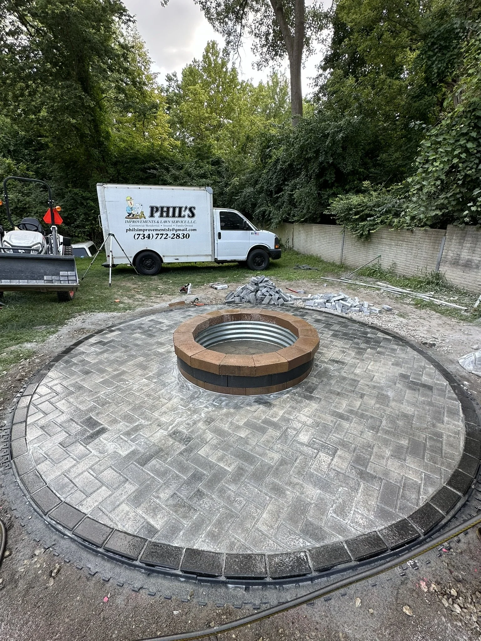 Construction site with a circular paved area and a central fire pit or structure, surrounded by tools, materials, and a white company truck with the logo 'Phil's Improvements & Lawn Service LLC' parked nearby, with trees and a wooden fence in the bac
