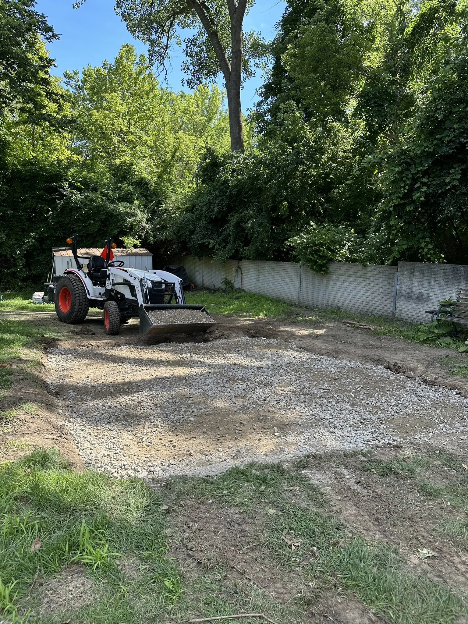 A small white tractor with red wheels parked on a dirt area with gravel, surrounded by green trees and bushes, with a metal fence in the background and a blue sky overhead.