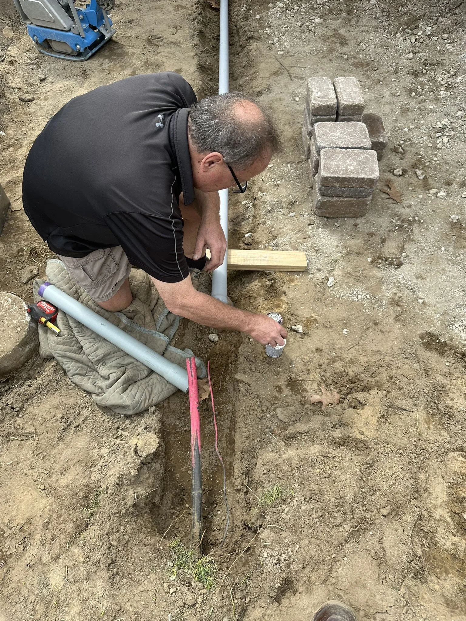 A man kneeling on the ground working on underground plumbing pipes, with a pipe, bricks, and tools nearby.