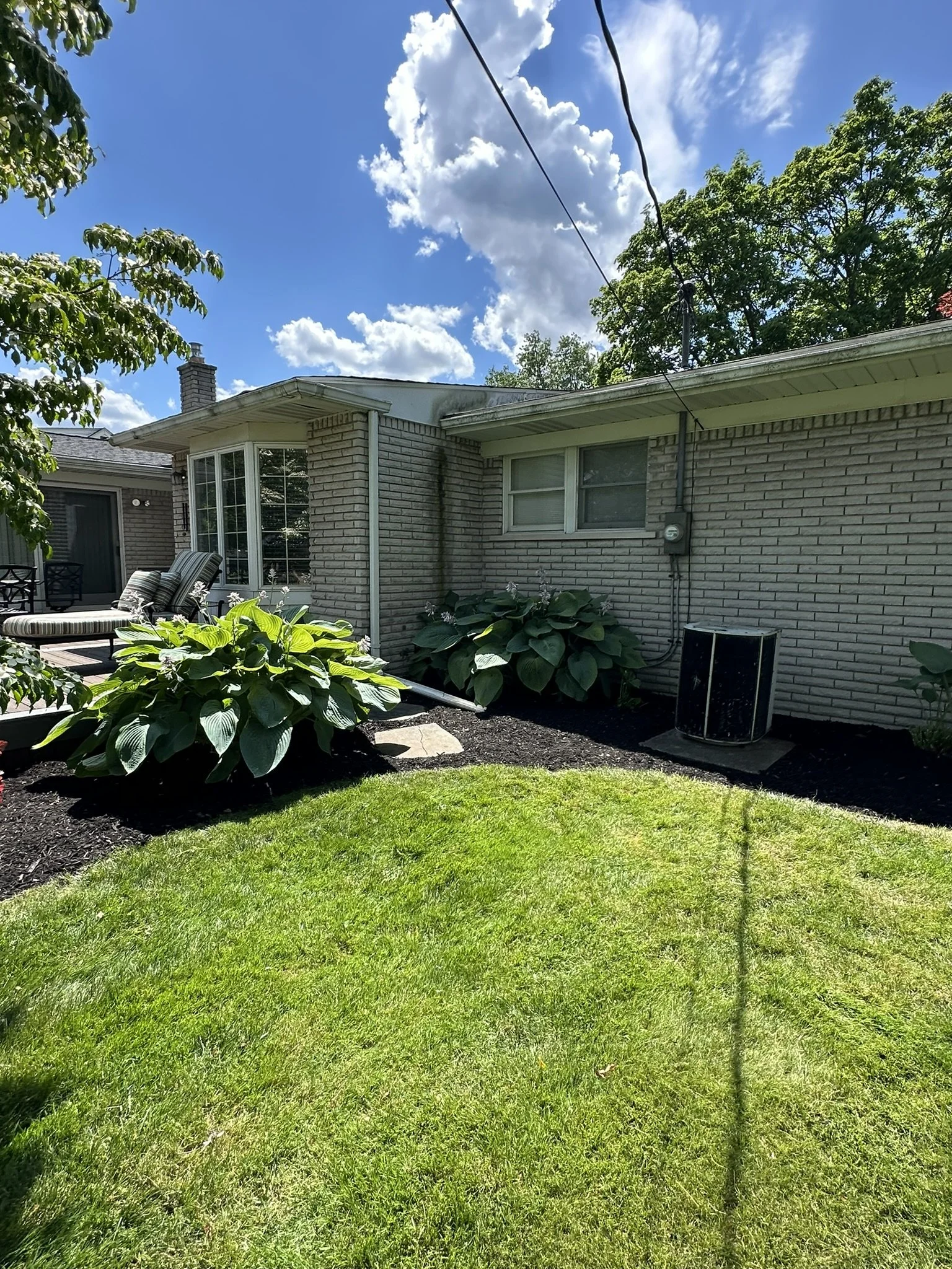 A backyard with green grass, shrubbery, outdoor chairs, and a brick house with windows. The sky is partly cloudy with sunlight.
