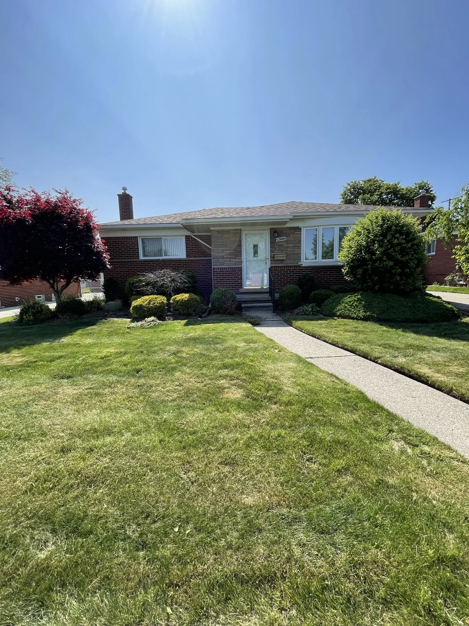 A single-story brick house with a front porch, surrounded by well-maintained lawn and shrubs, under a clear blue sky.