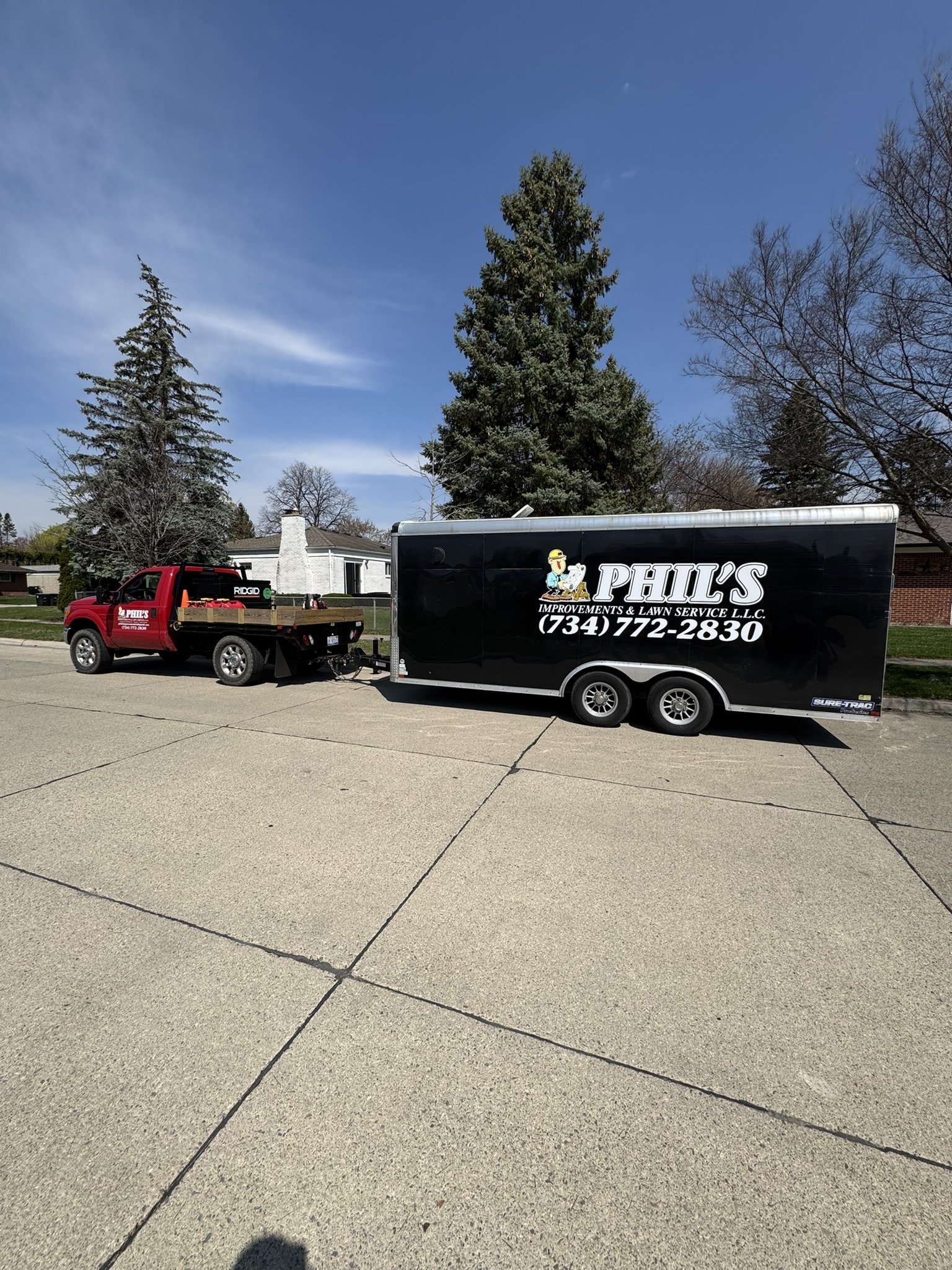 A red pickup truck towing a black trailer with white text that reads 'Phil's Improvements & Lawns Service LLC' and a phone number. The scene is on a residential street with trees and a house in the background under a blue sky.