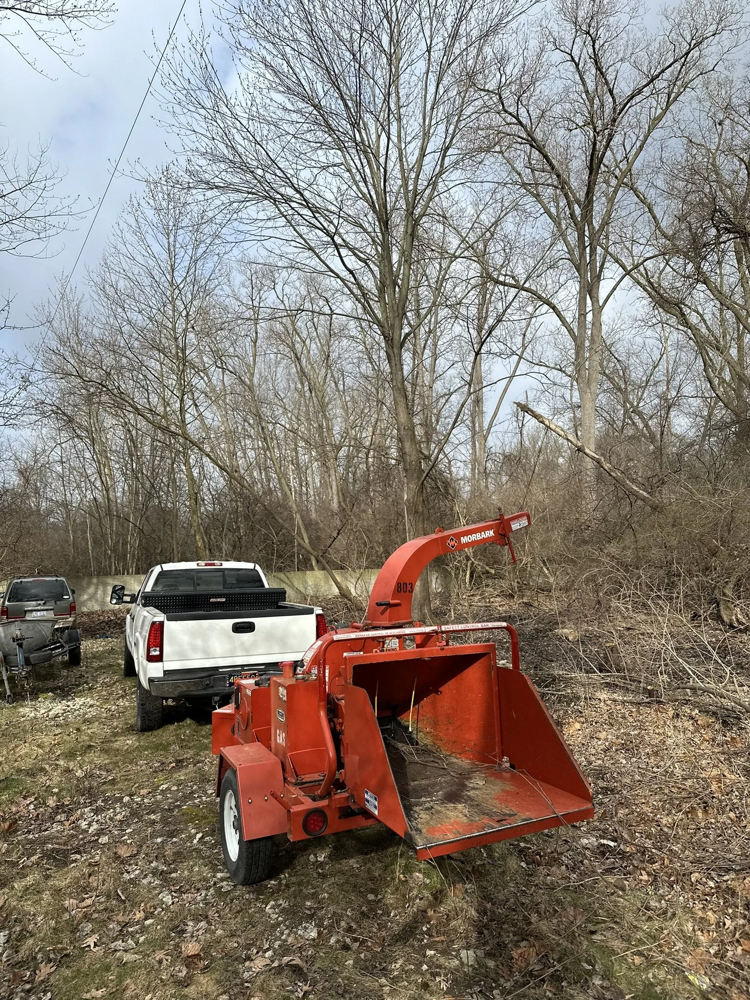 A red wood chipper machine parked outdoors on a grassy area surrounded by leafless trees and vehicles, including a white truck and a black SUV.