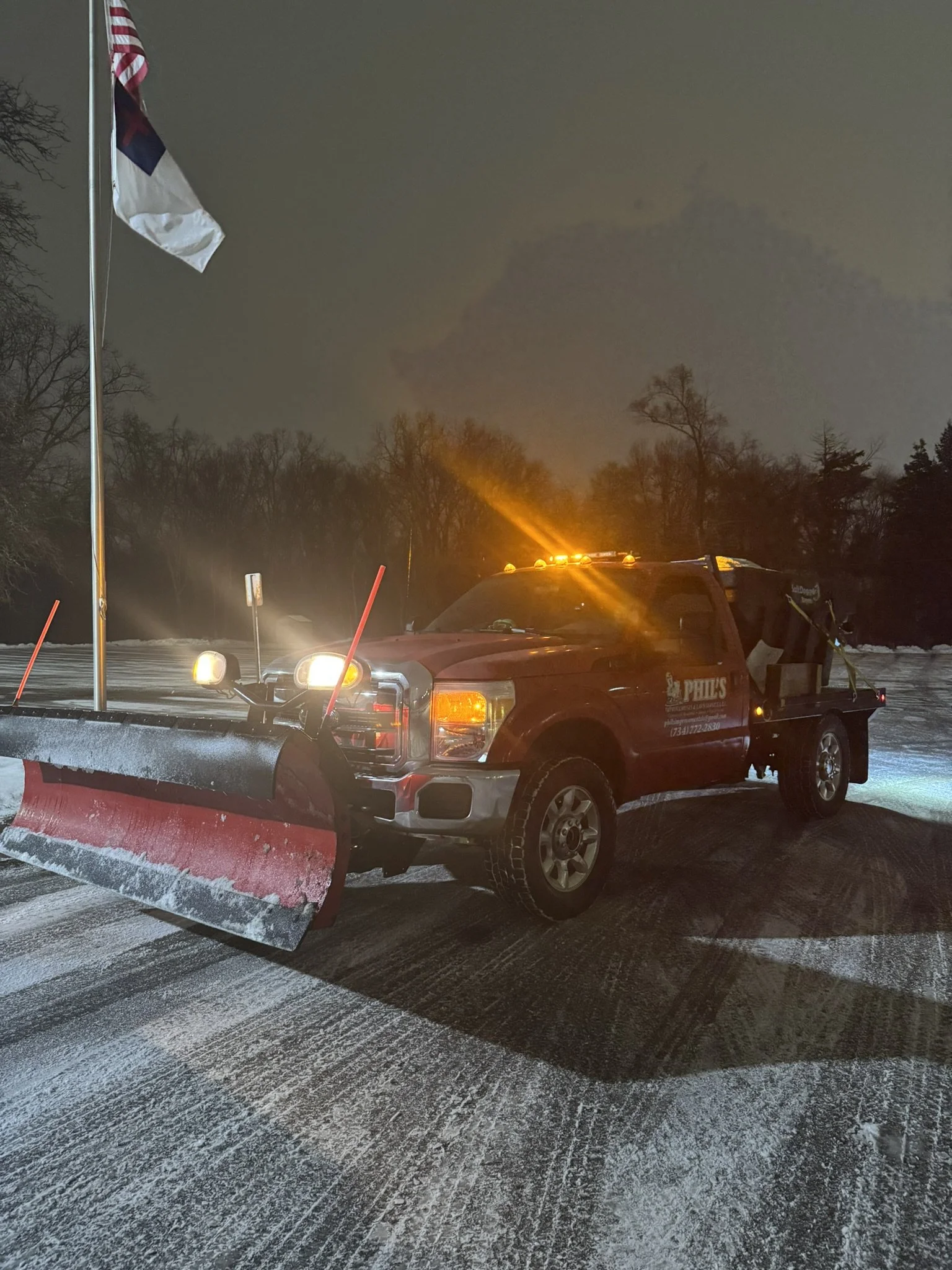 A red work truck with a snow plow attached is parked on a snowy road at night, with orange flashing lights on top and a flagpole with a flag nearby.