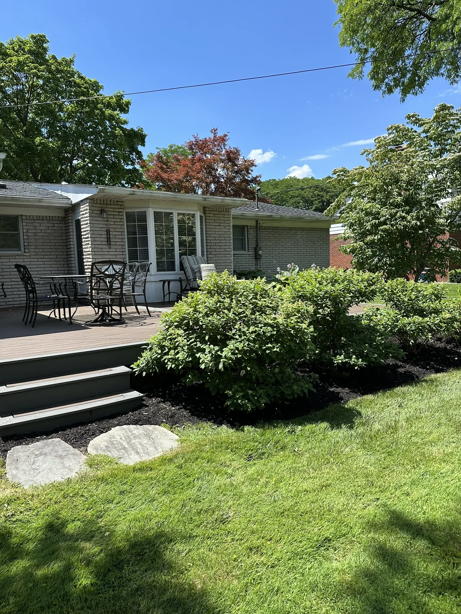 A backyard with a wooden deck, black metal chairs, and a table. There are lush green bushes, a tree with green and reddish leaves, and a grassy lawn. The house is made of brick with a bay window, and the sky is blue with some clouds.