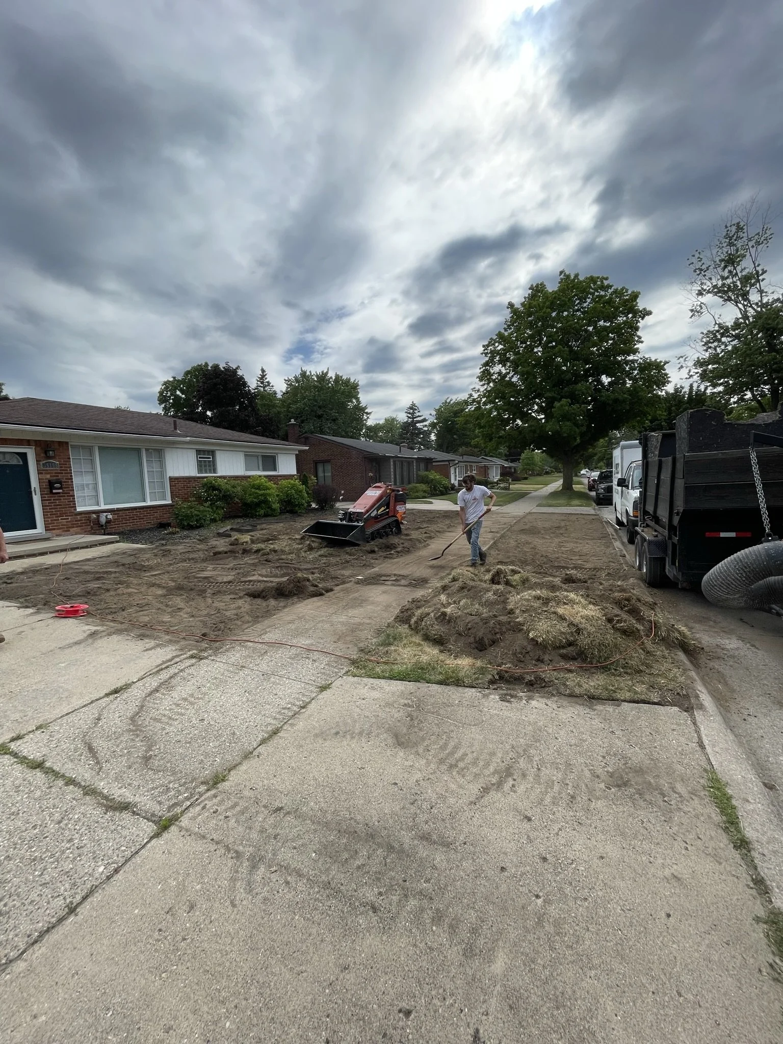 People working on residential sidewalk repair with landscaping equipment, dirt mounds, and trucks, under cloudy skies in a suburban neighborhood.