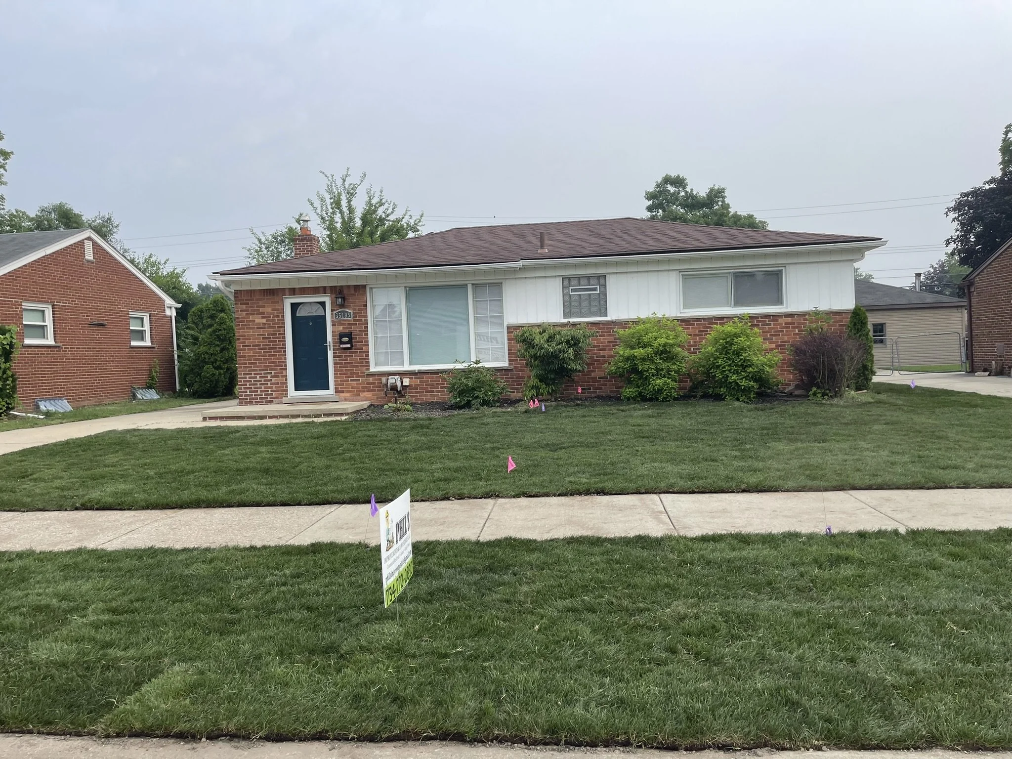 A house with brick facade and white siding, blue front door, front yard with green grass, bushes, and a sidewalk.