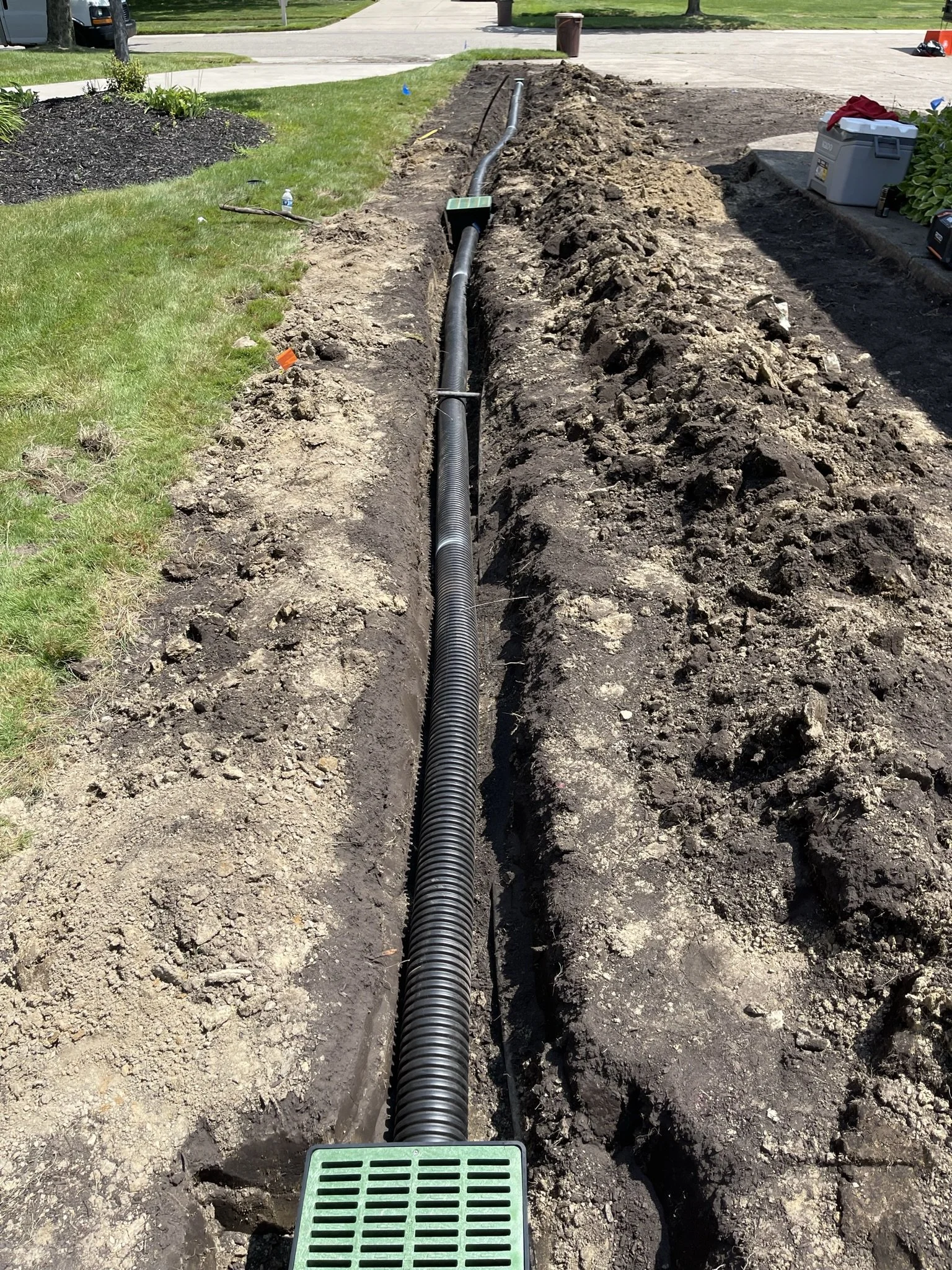 Installation of underground conduit or cable in a trench, with a drainage grate in the foreground and a toolbox nearby.