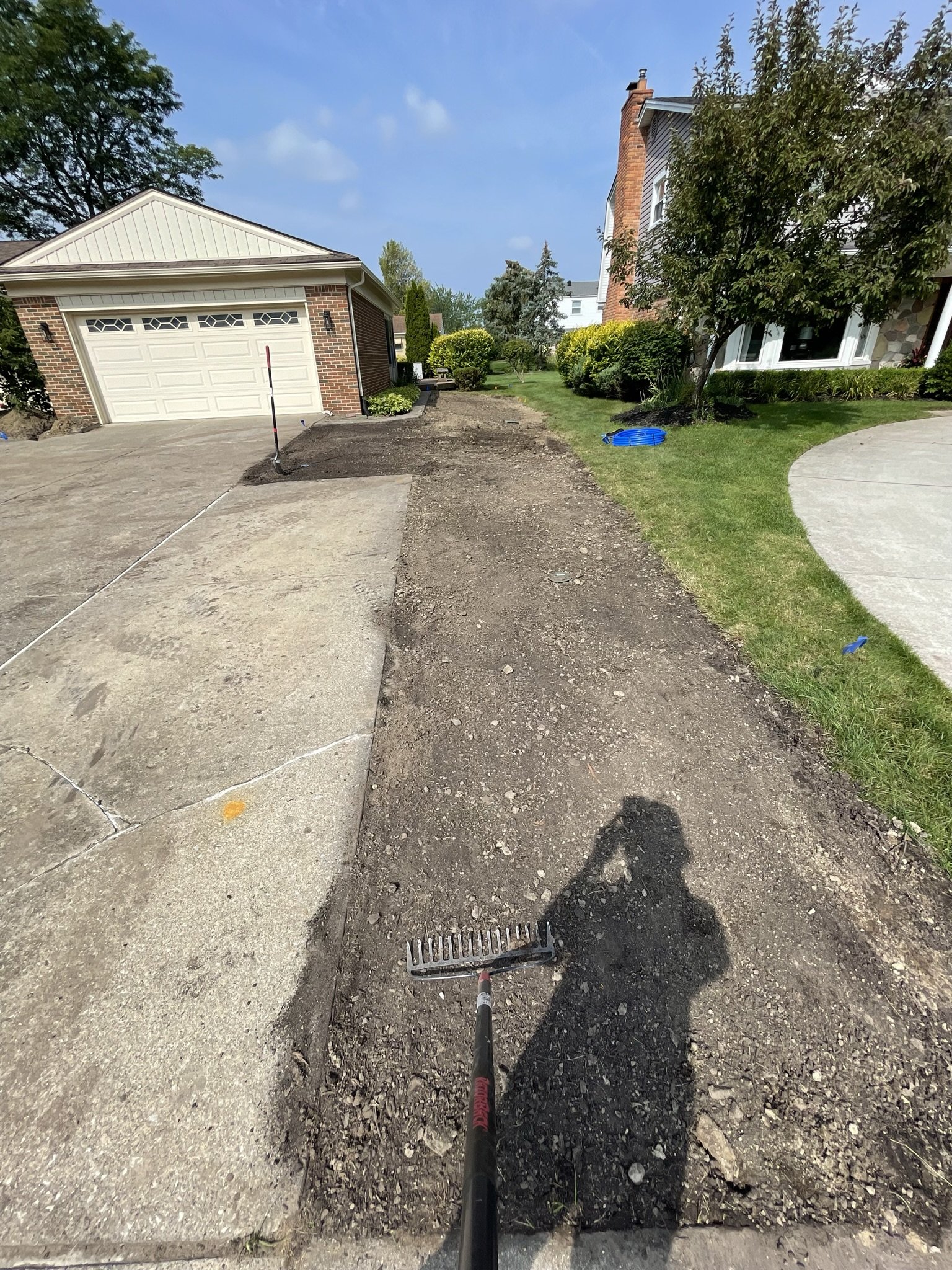 A residential front yard with an ongoing sidewalk or pathway repair, showing a person holding a rake, with part of the concrete driveway on the left and a grassy lawn with bushes and trees on the right, under a mostly sunny sky.