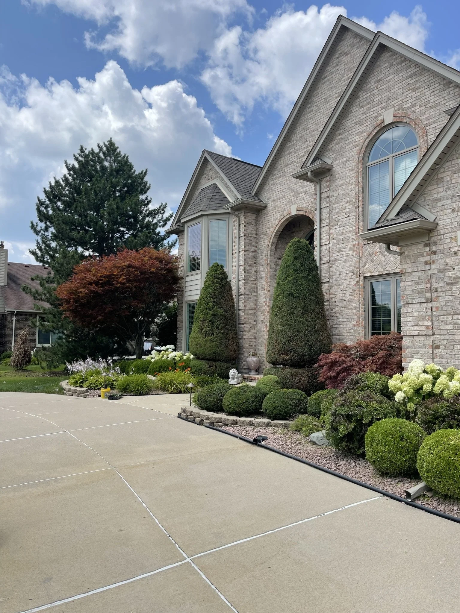 Front yard of a large brick house with manicured bushes and trees, a driveway, and a partly cloudy sky.