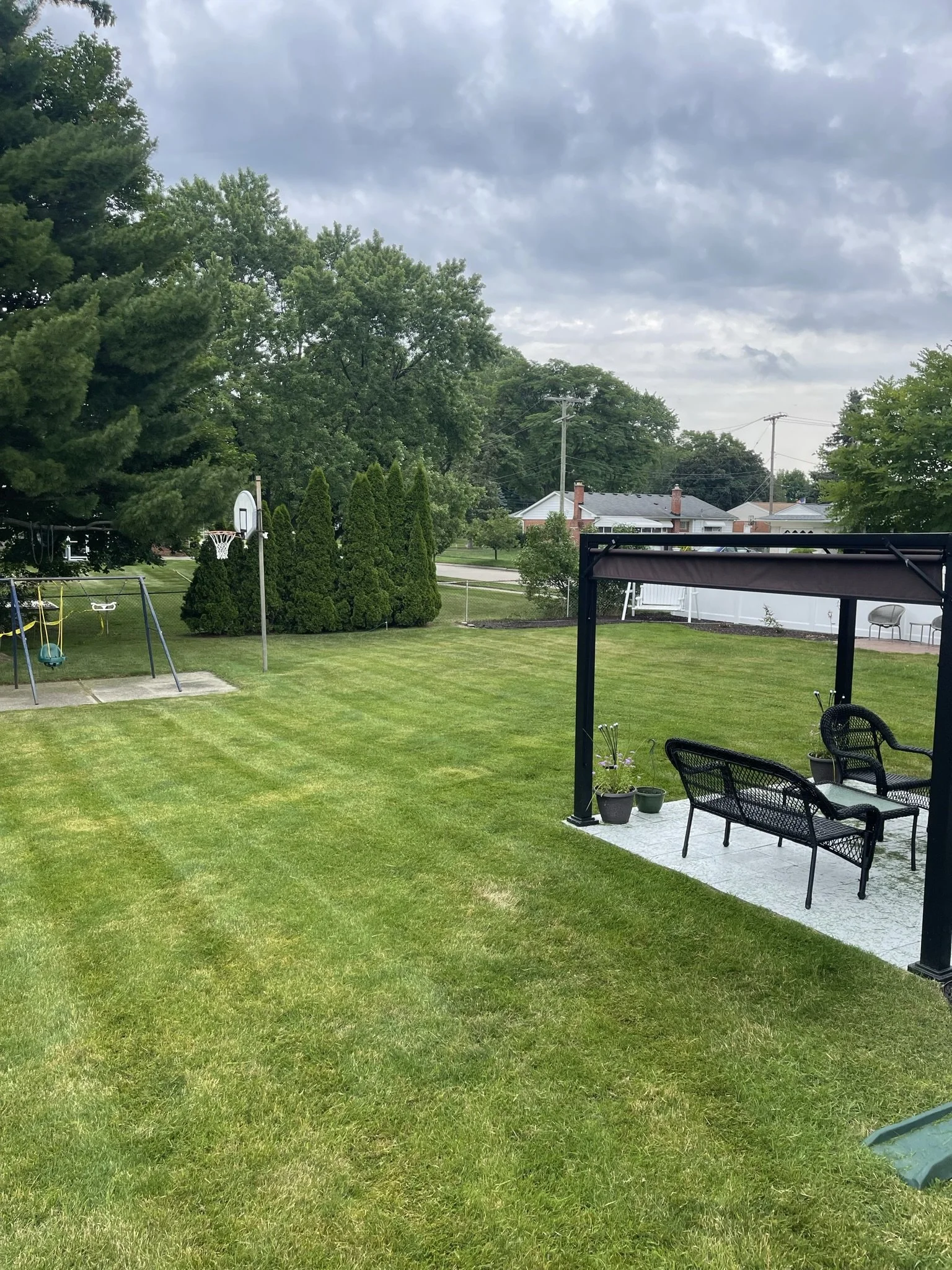 A backyard with a well-maintained grassy lawn, a swing set and basketball hoop on the left, a pergola with outdoor furniture and potted plants on the right, trees, and neighboring houses and power lines in the background under a cloudy sky.