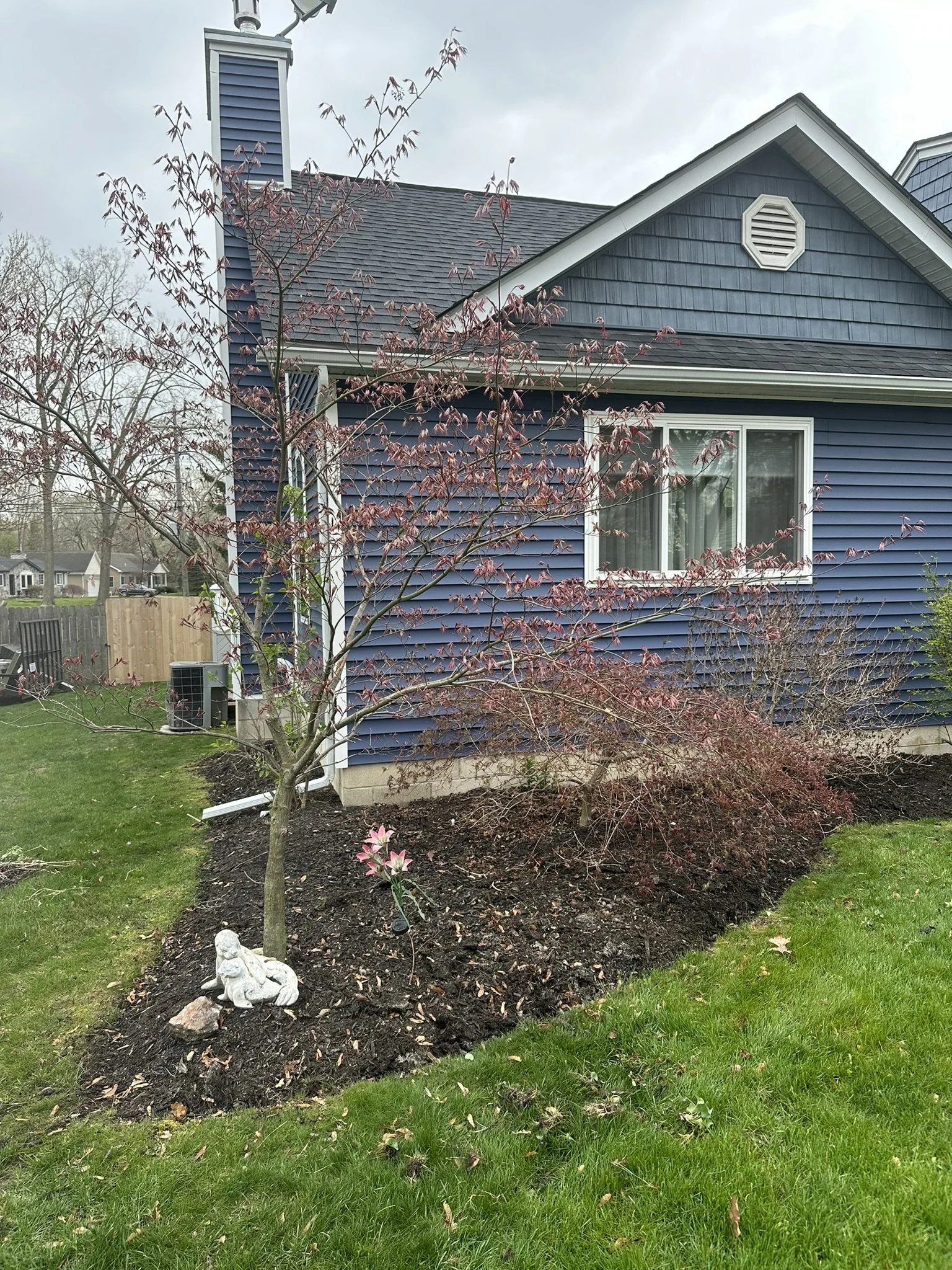 A blue house with white trim and a window, a small tree with reddish leaves, a garden bed with a moss-covered stone and a white garden gnome, a young flowering plant, and a wooden fence in the background.