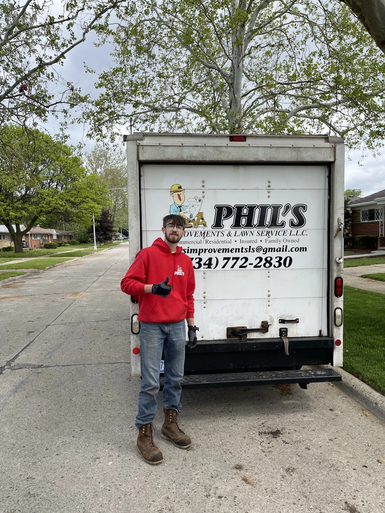 A man wearing a red hoodie, jeans, and work boots standing in front of a white truck with the business logo for Phil's Improvements & Lawn Service, giving a thumbs-up.