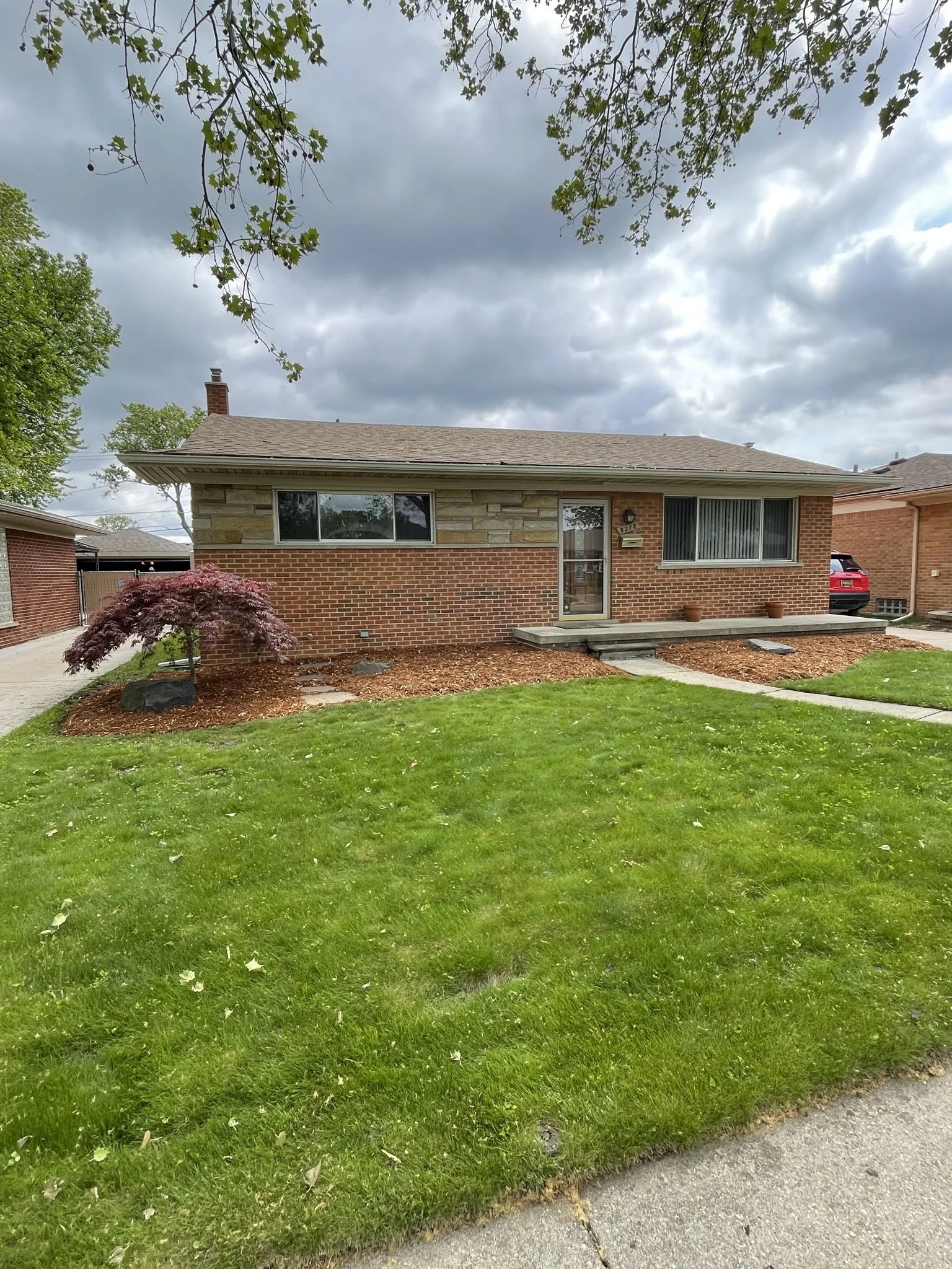 Split-level house with brick and stone exterior, large front window, small porch with steps, and well-maintained lawn with small trees and shrubs; cloudy sky overhead.