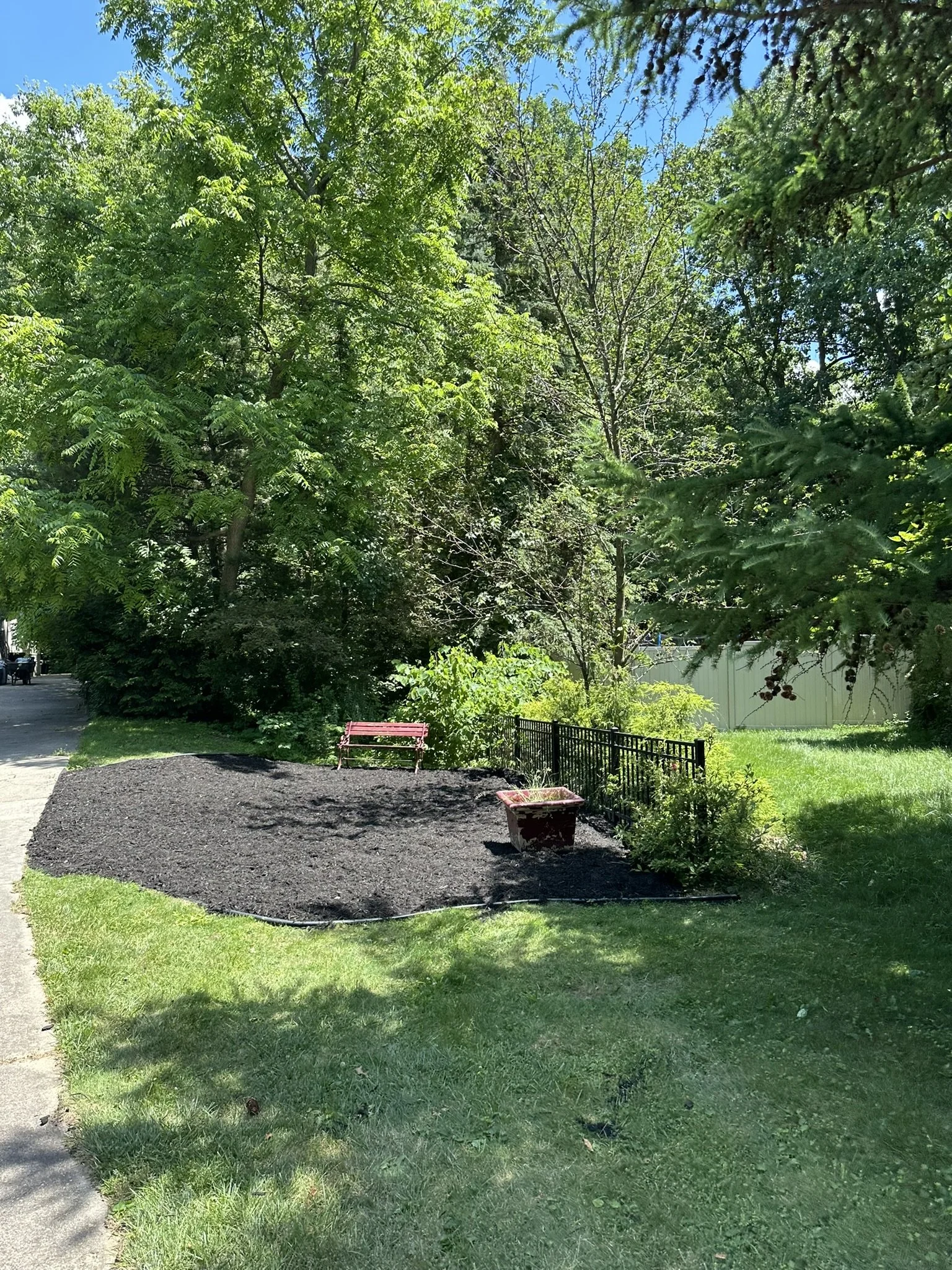A small, freshly mulched garden bed with a pink bench, a planter, and a black fence, surrounded by green grass and trees on a sunny day.