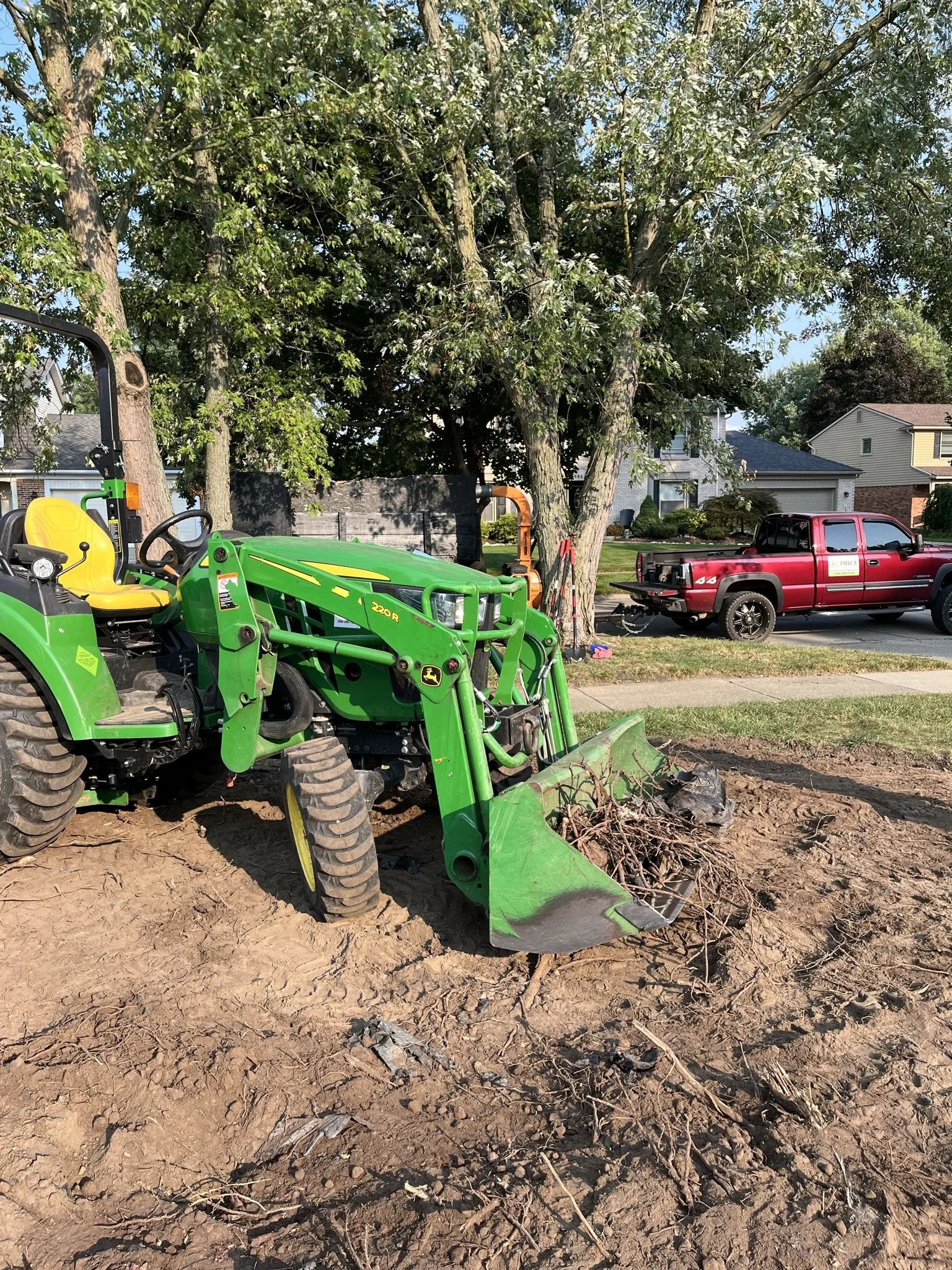 A green John Deere tractor with a front loader attachment filled with branches and debris, parked on bare soil next to a sidewalk, with a large tree and suburban houses in the background.