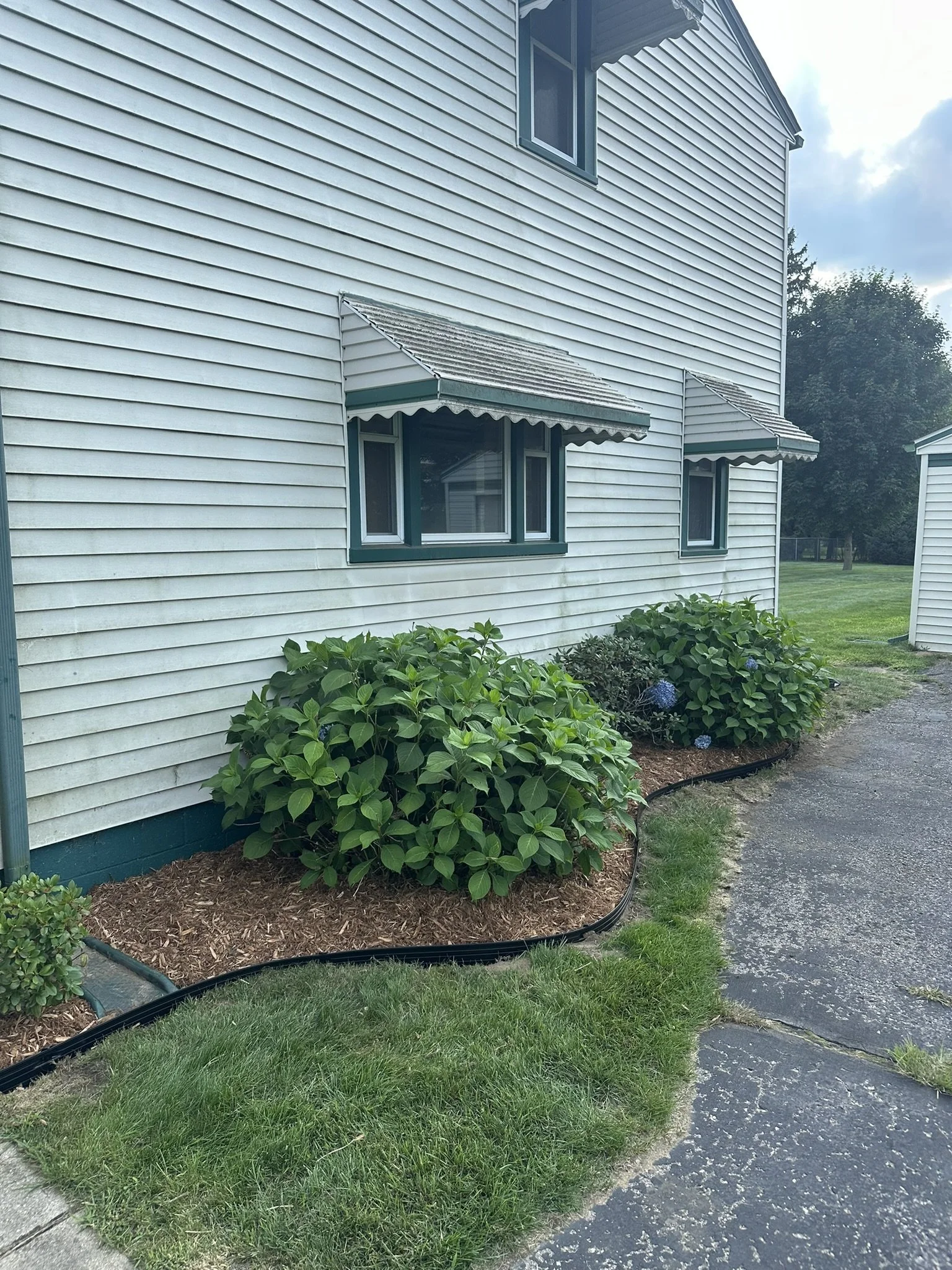 Side of a white house with three small windows, each with a green frame and a green and white striped awning. There are well-kept bushes with purple flowers in front of the house, landscaped with mulch and a black edging. A gravel driveway runs along