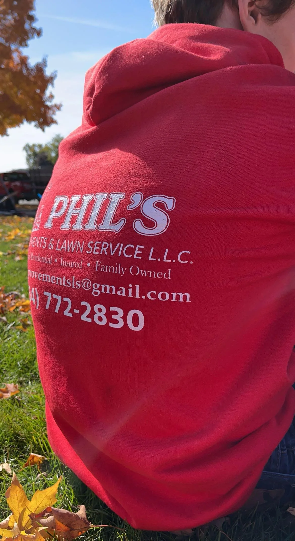 Close-up of a person wearing a red hoodie with white text advertising a lawn and landscape service, including contact information, outdoors with grass and autumn leaves.