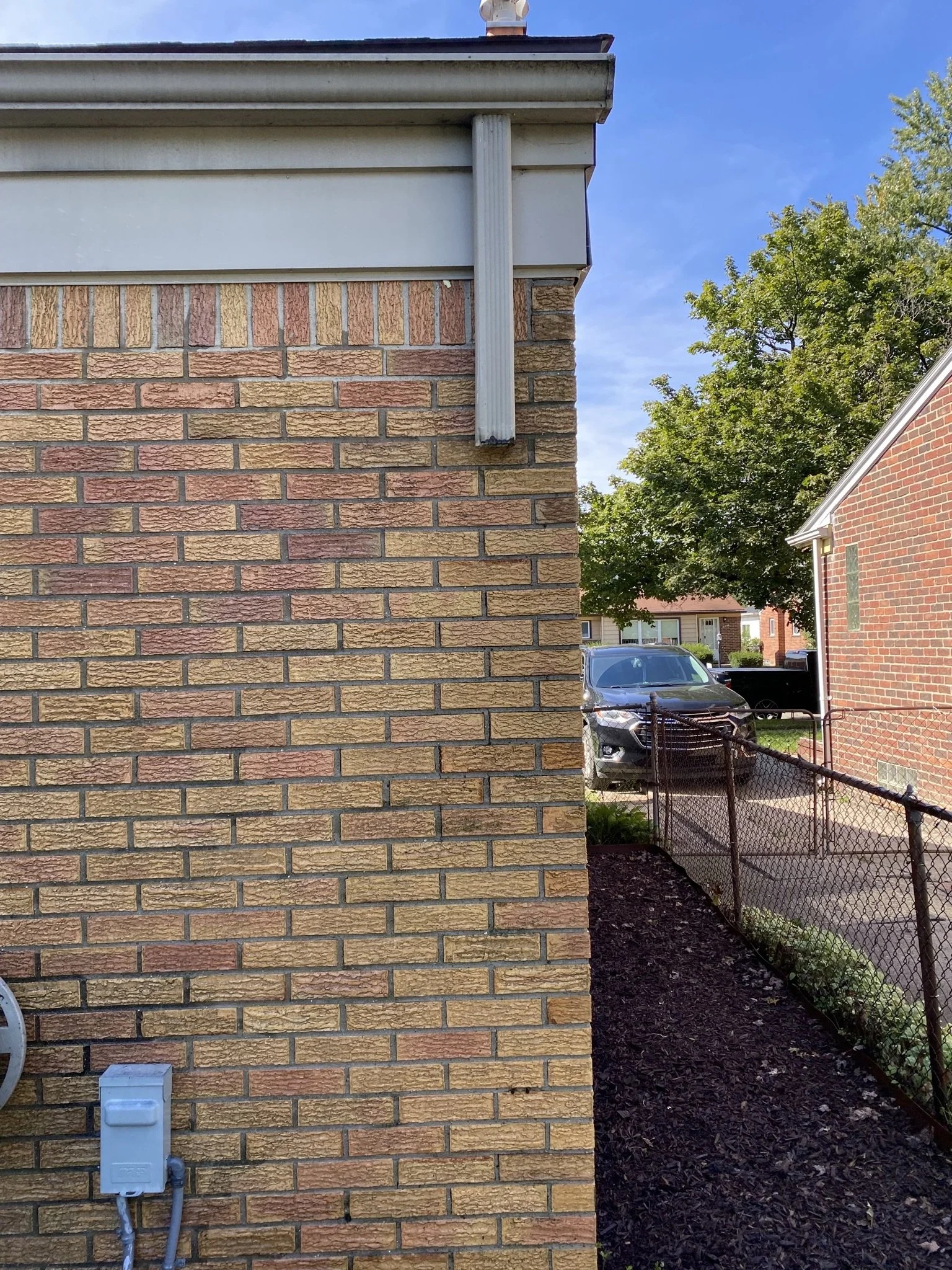 Side view of a brick building with a corner and a chain-link fence, a parked black SUV, trees, and a blue sky in the background.