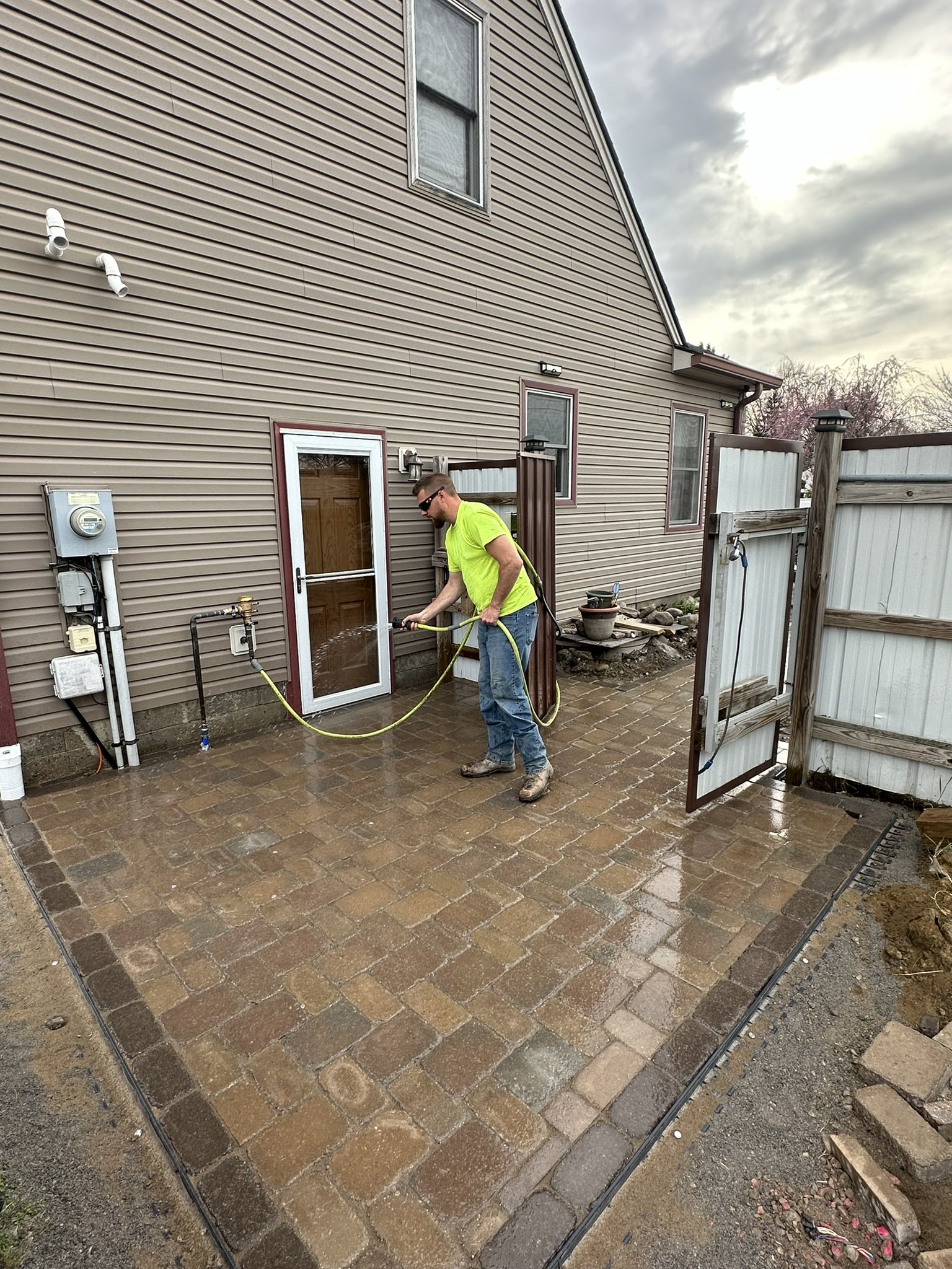 Man pressure washing a brick patio outside a beige house with white trim on a cloudy day.