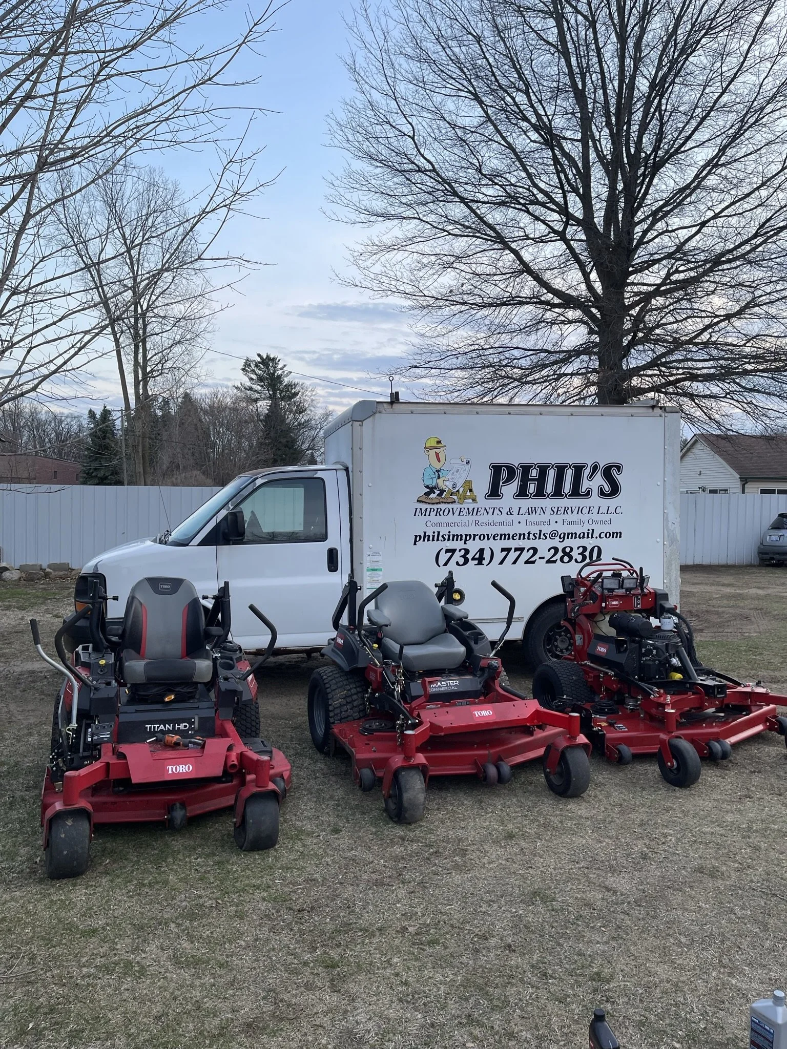 Three red commercial lawn mowers parked on grass in front of a white truck with business information for Phil's Improvements & Lawn Service, and large trees in the background.