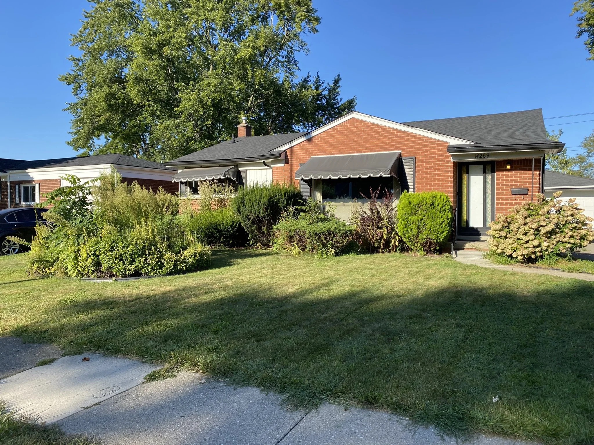 A single-story brick house with a black roof and front porch, surrounded by shrubs and a well-maintained lawn, under a clear blue sky.