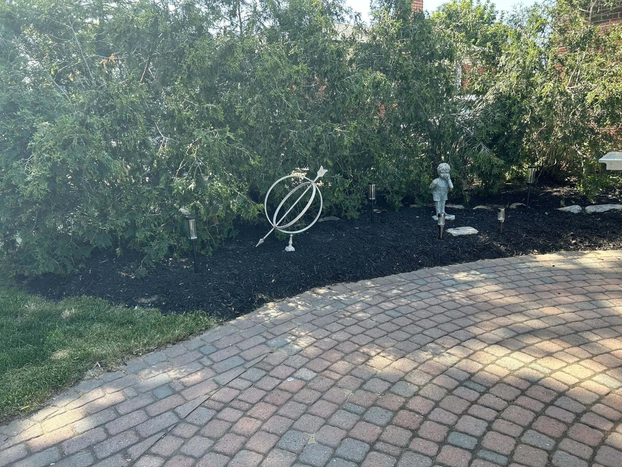 Garden with small statue of a girl, decorative metal ball, and multiple small pathway lights, mulch soil, next to brick patio and green shrubbery.