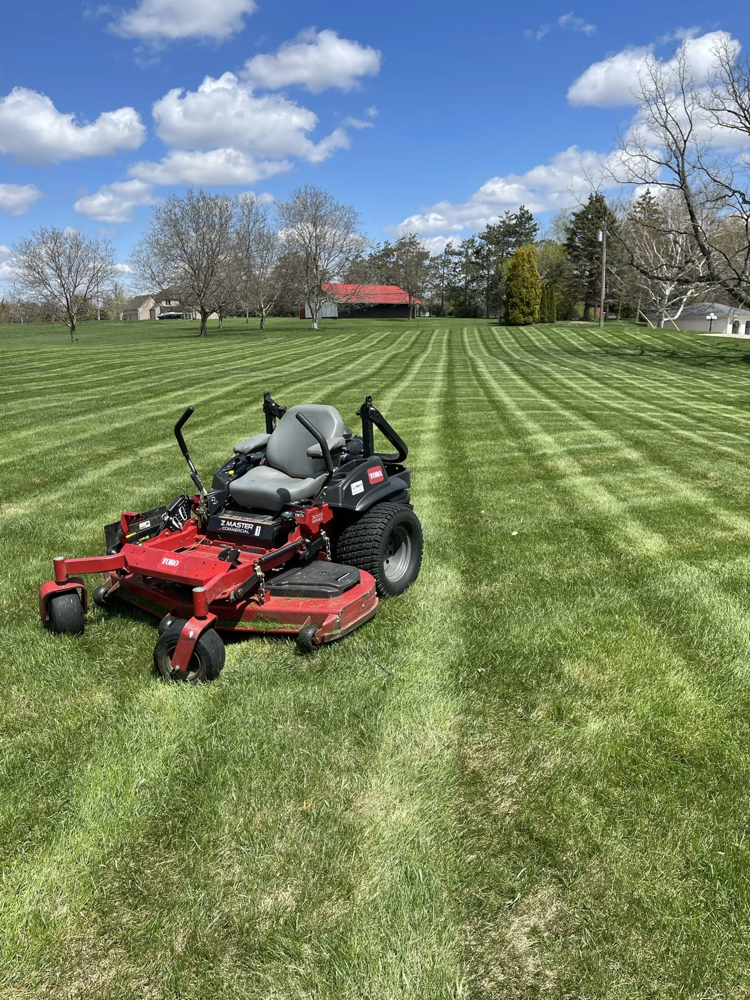 A red and black zero-turn lawn mower on a freshly mowed grassy field under a partly cloudy blue sky.