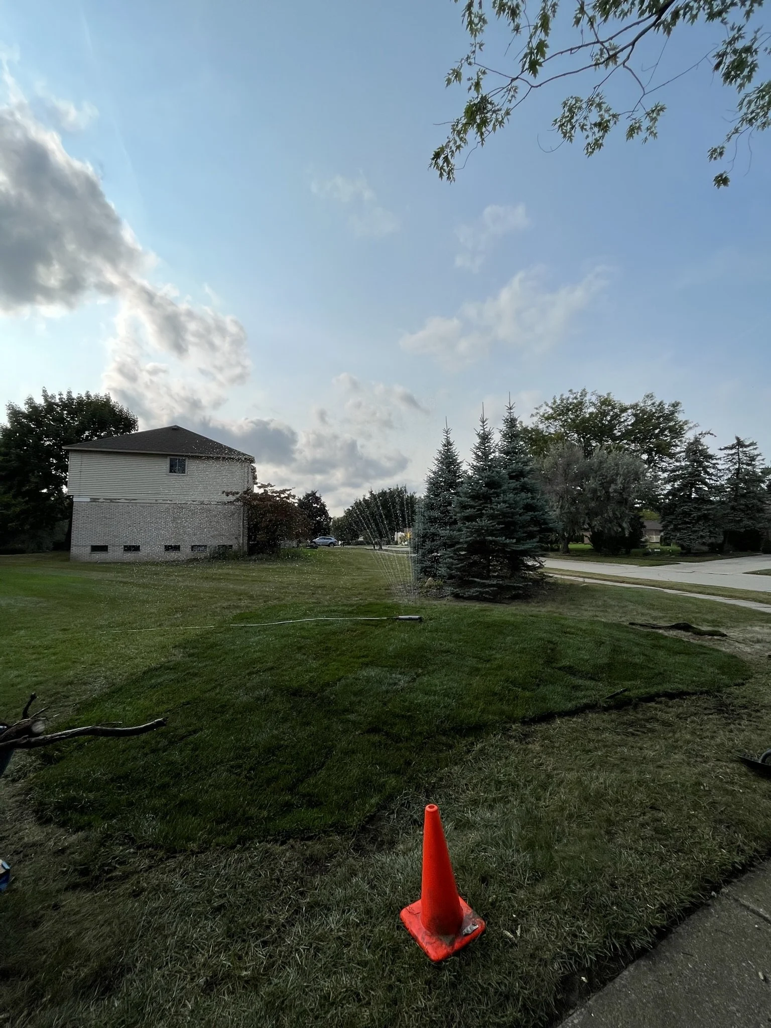 A lawn being watered with a sprinkler, with orange traffic cone in the foreground, a house and trees in the background, and a partly cloudy sky overhead.