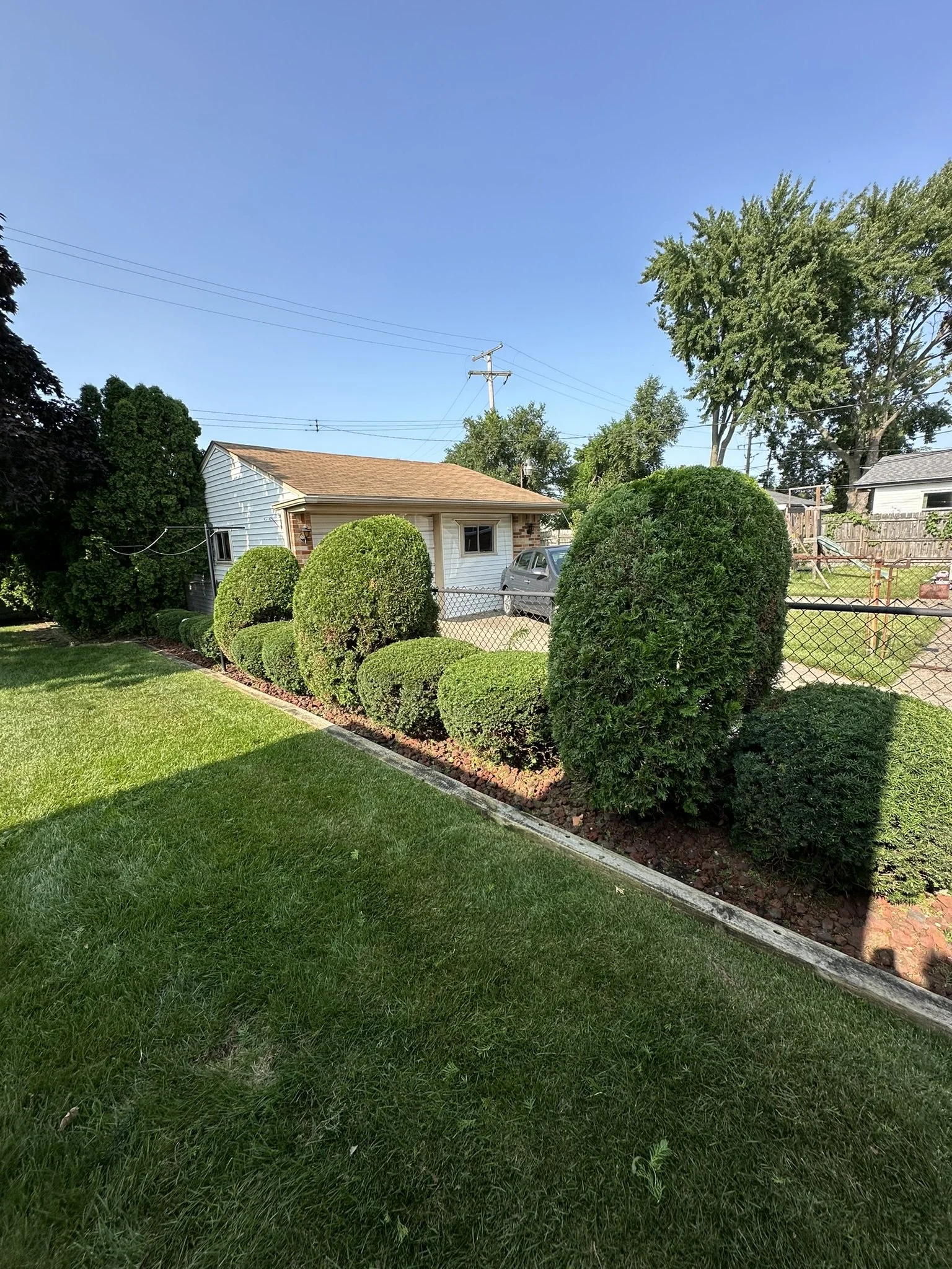 Well-manicured front yard with neatly trimmed bushes along the fence, a small house in the background, and a clear blue sky.