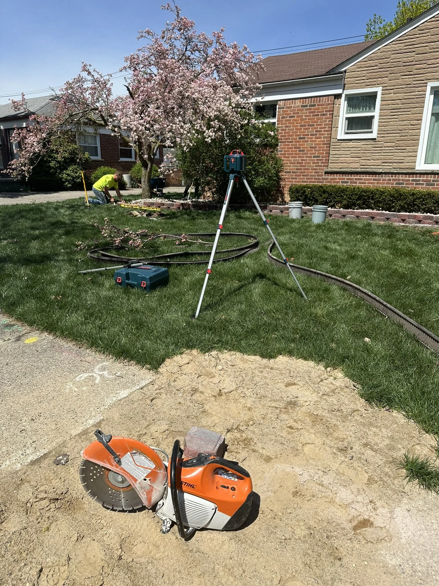 A construction site in a residential yard with a cherry blossom tree, a worker in a yellow shirt working in soil, a tripod-level surveying instrument, a lasered device on the ground, and a portable saw in the foreground. There are houses in the backg