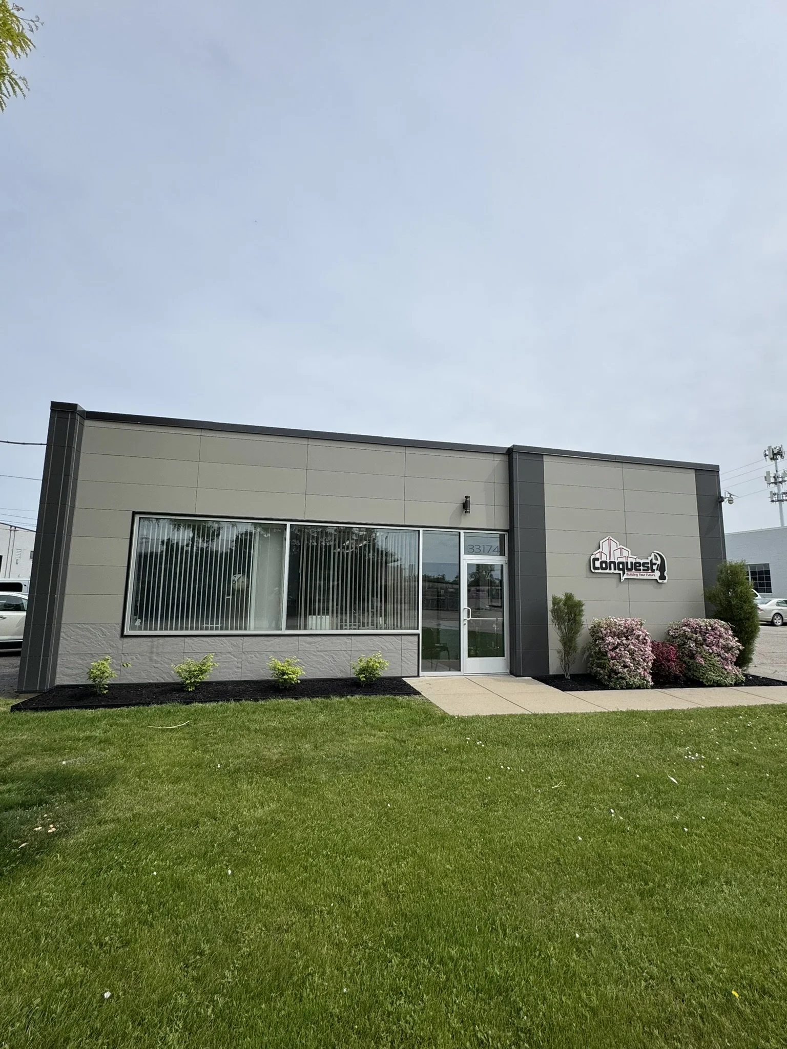 Exterior of a commercial building with a sign that reads 'Conquest.' The building has a large window with blinds, a glass door, and landscaped bushes and grass in front.