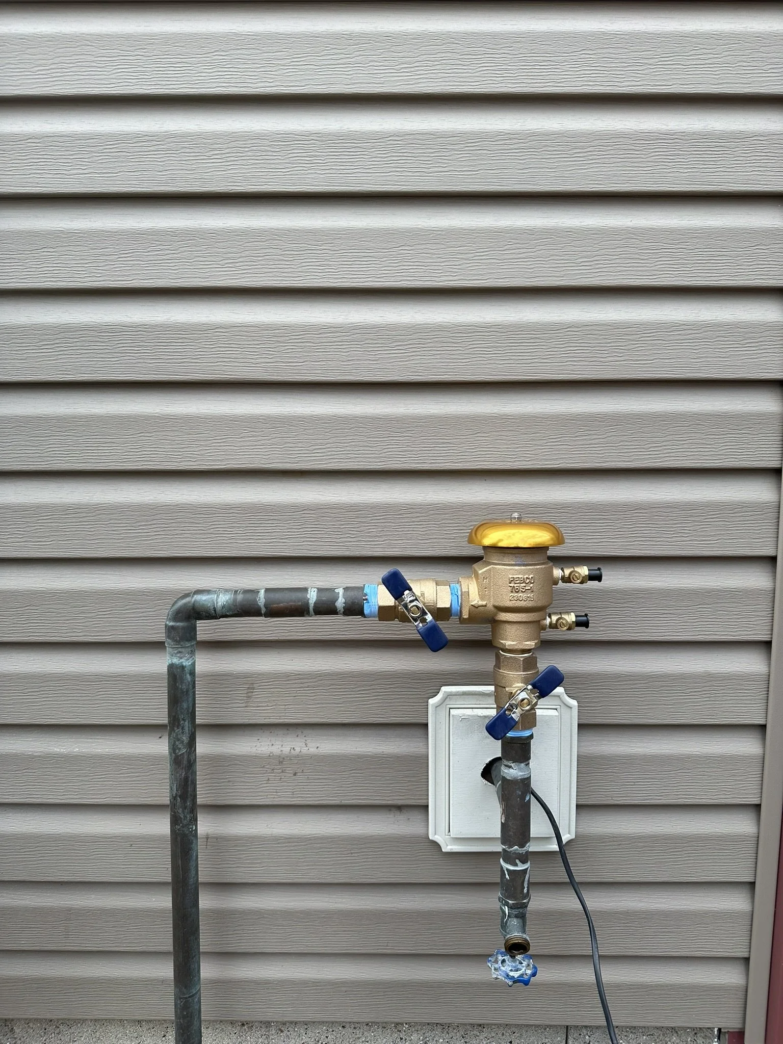 A gas meter attached to the side of a beige vinyl siding house, with piping, valves, and a power cord connected to it.