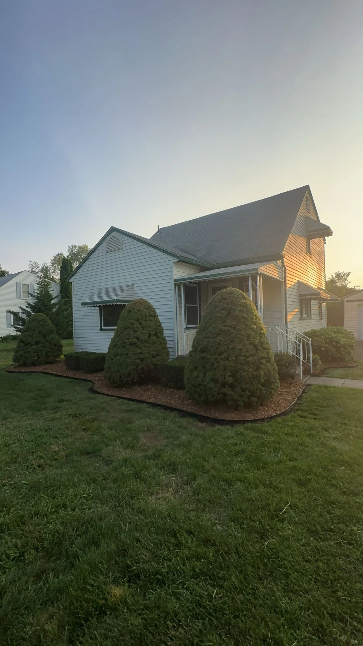 A white house with a sloped roof and small porch, surrounded by neatly trimmed bushes and a green lawn during sunset.