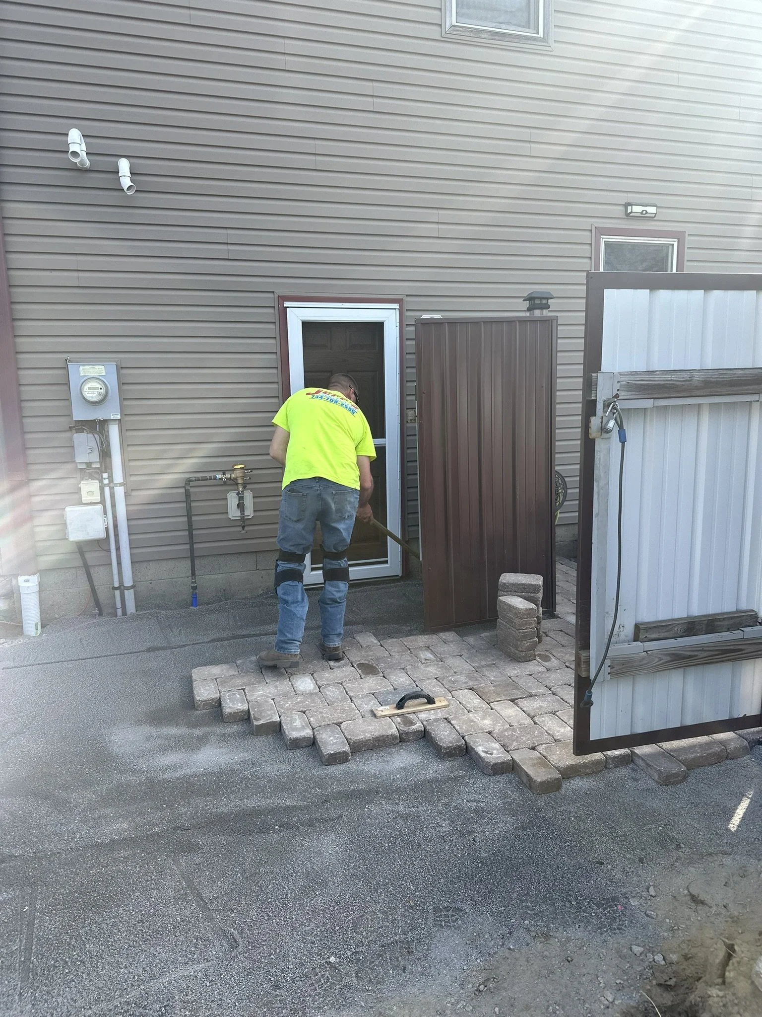 A worker installing pavers outside a house, with a clipboard on the ground nearby.
