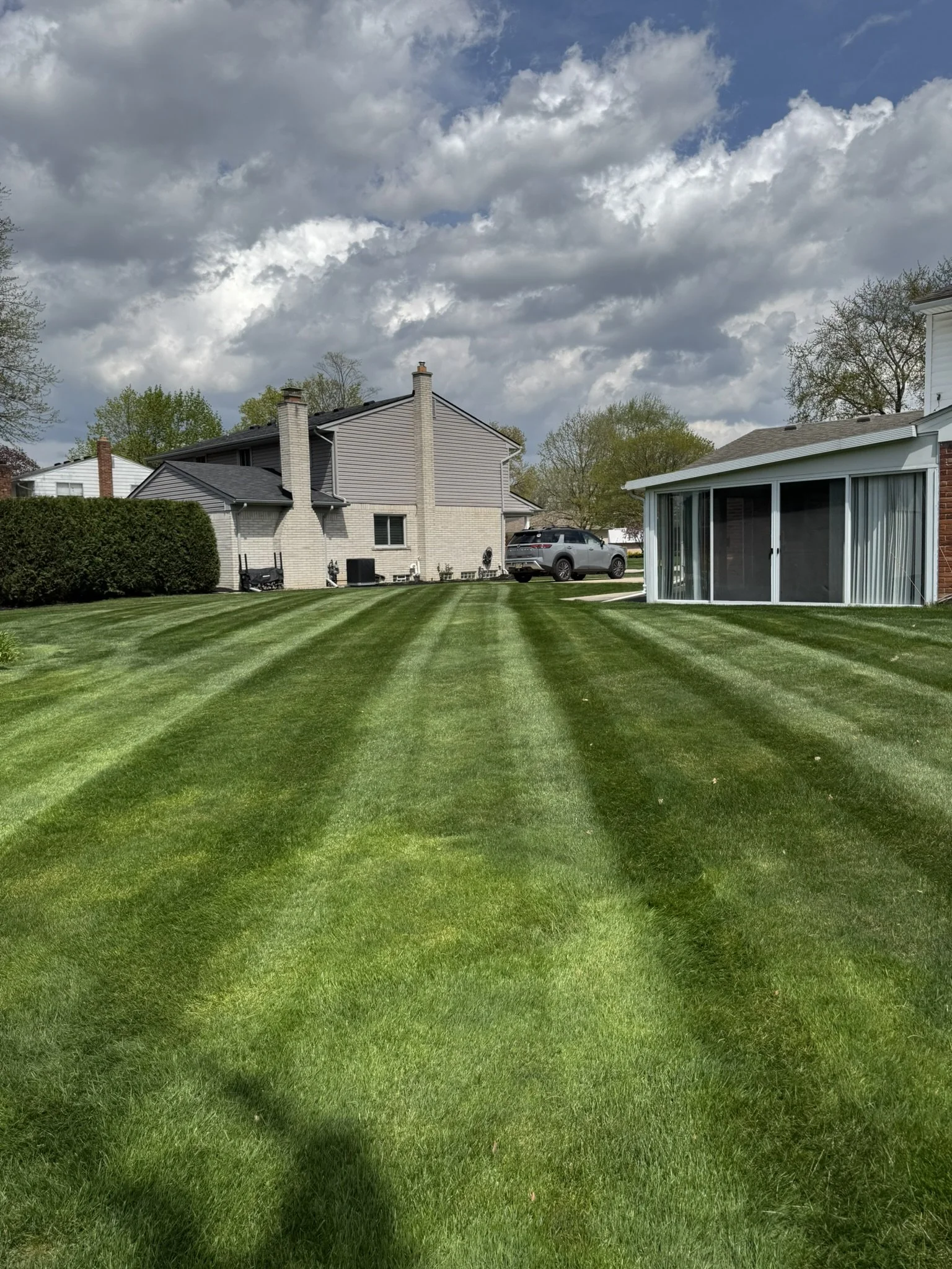 Freshly mowed backyard with green grass and striped pattern, white brick house with car parked in driveway, screened porch on the right, blue sky with clouds.