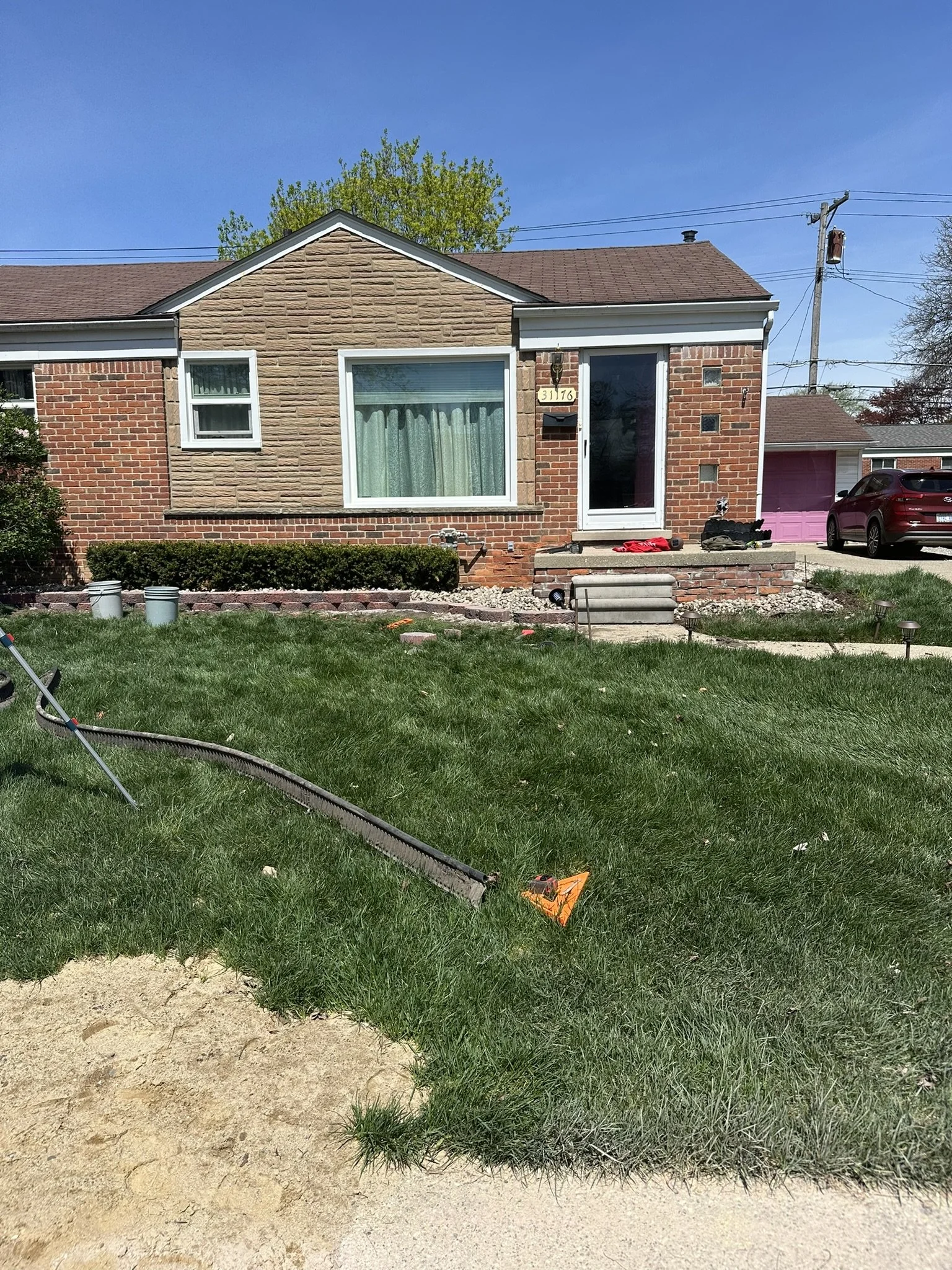 A single-story brick house with a large front window, green curtains, and a small porch with steps. There is a well-kept lawn with some tools and equipment on the grass, and a driveway with a pink garage door and a red car parked nearby. Power lines 