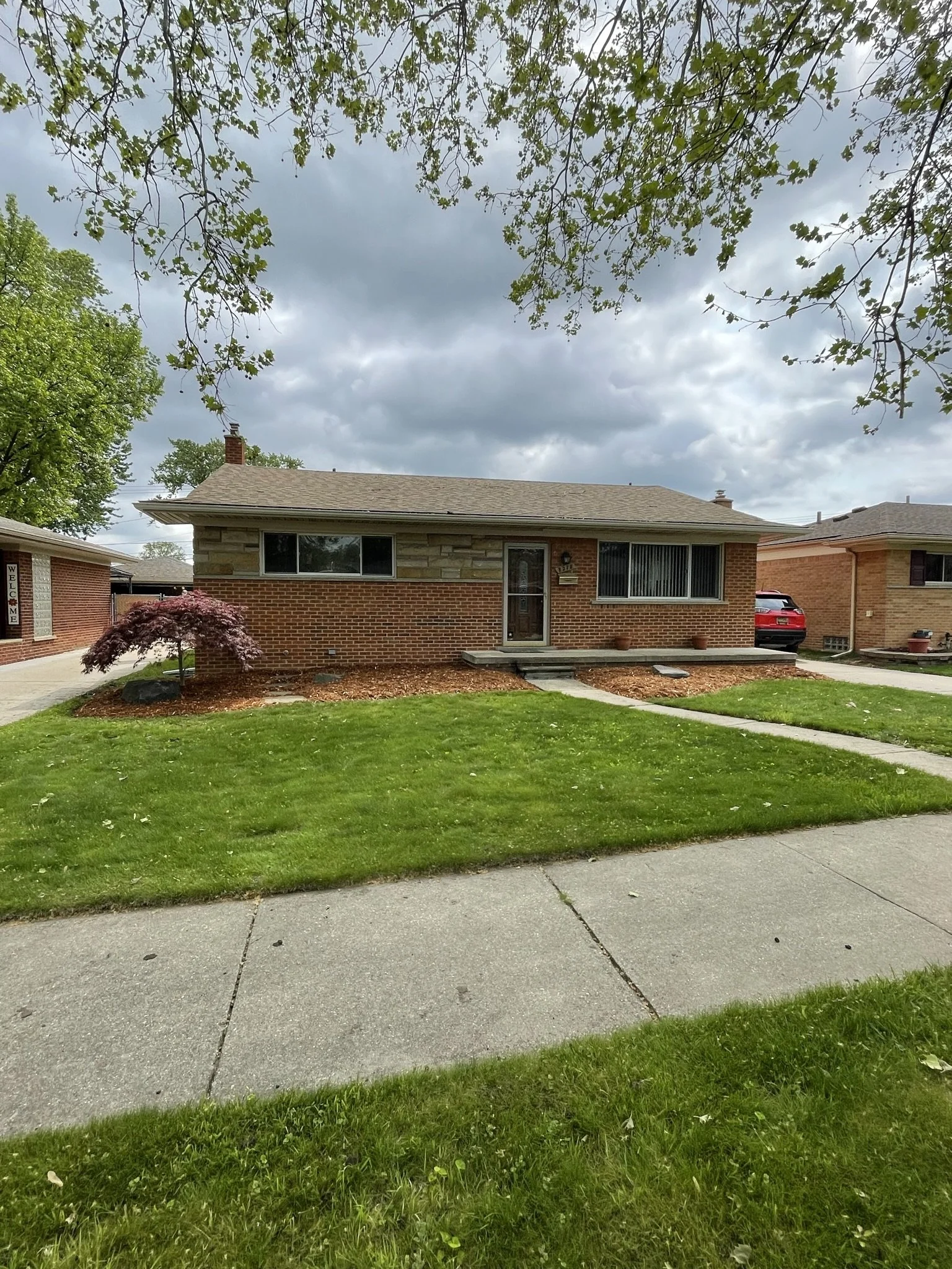 A single-story brick house with a brown shingle roof, concrete steps leading to the front door, a small landscaped front yard with green grass and a purple-leafed tree, and a driveway with a parked red car, under a partly cloudy sky.