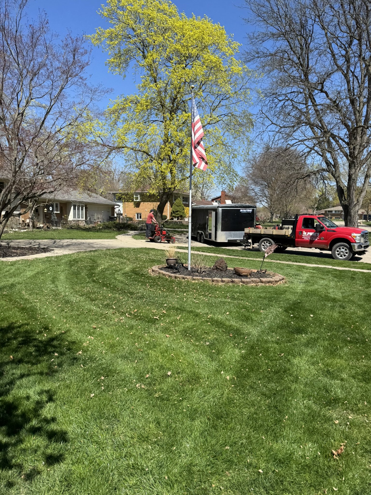 A residential front yard with a green lawn, a large tree with yellow-green leaves, an American flag on a pole, a person mowing the lawn, and a red truck attached to a black trailer parked on the driveway. There are houses in the background and a clea