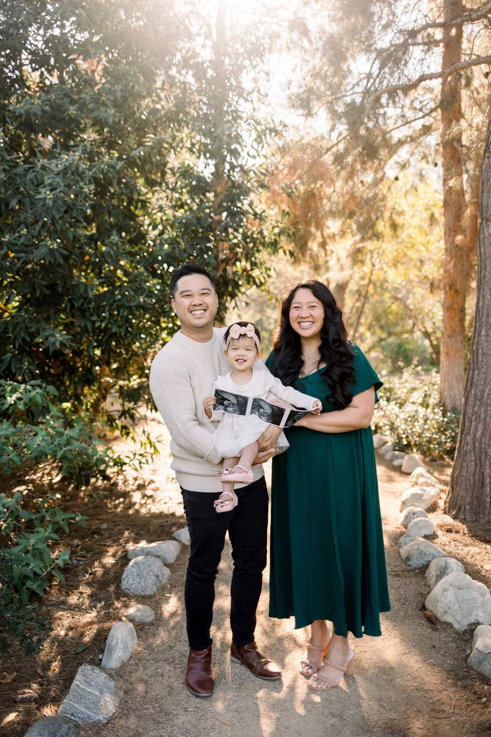 A family of three standing in a park trail, smiling with an ultrasound photo, announcing their pregnancy.