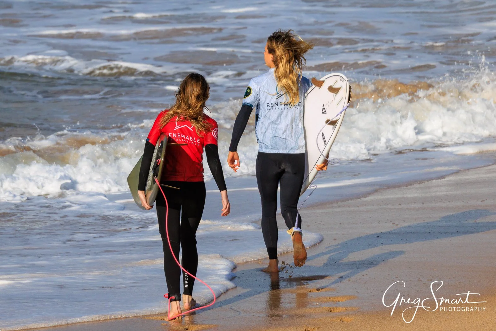 Two women walking on the beach carrying surfboards, one wearing a red wetsuit and the other a white wetsuit, with ocean waves in the background.