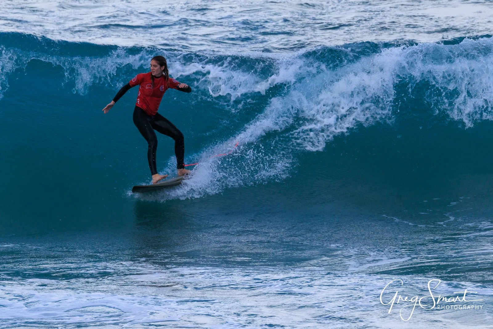 A woman surfing on a large blue wave, wearing a red and black wetsuit, with a focused expression.