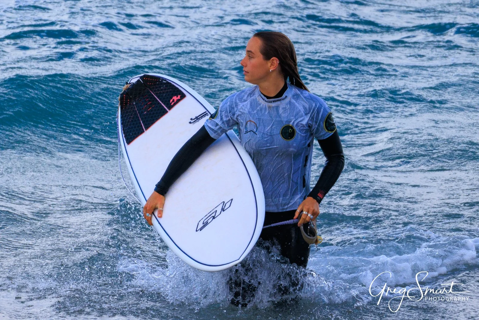A woman in a wetsuit holding a white surfboard, walking out of the ocean.