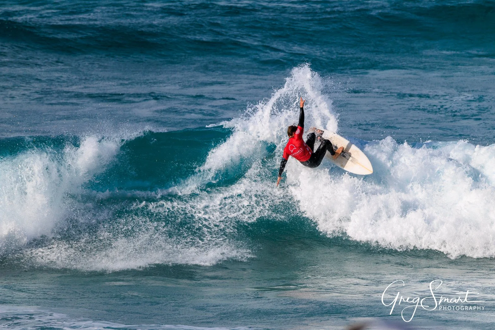 A surfer riding a wave in the ocean, wearing a red wetsuit and black surfboarder shorts, with one arm extended upwards and the other down, amidst white foamy surf.