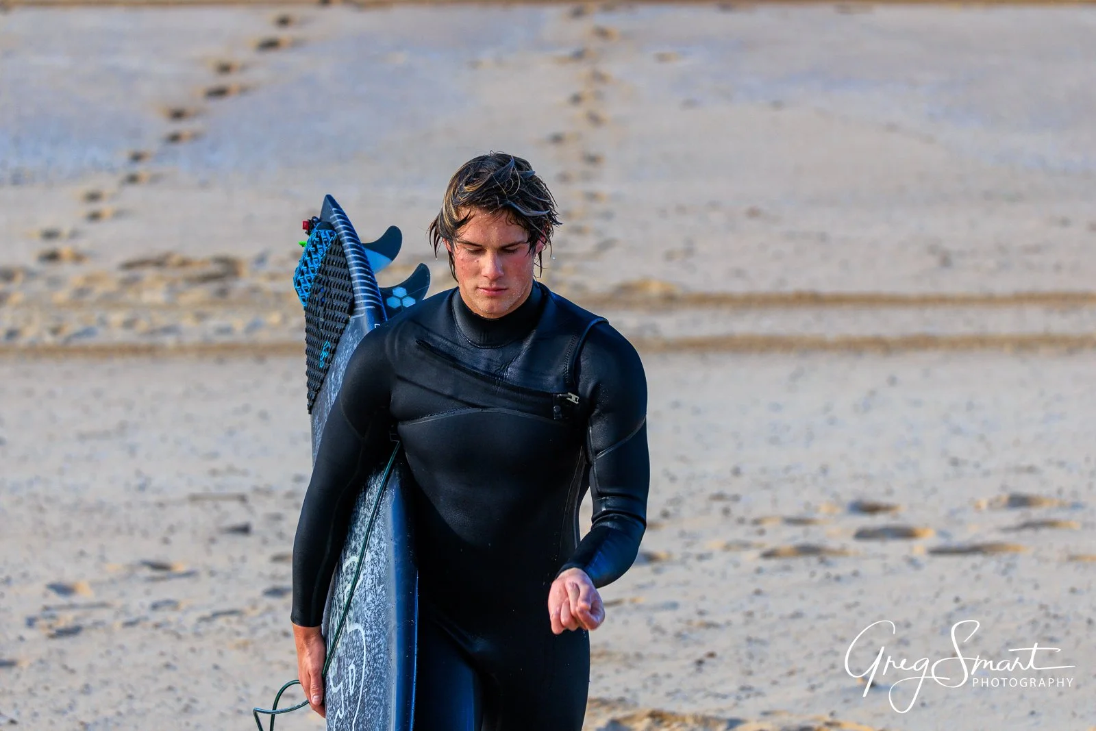 Young man in a black wetsuit carrying a surfboard on a sandy beach, looking down and walking away from the water.