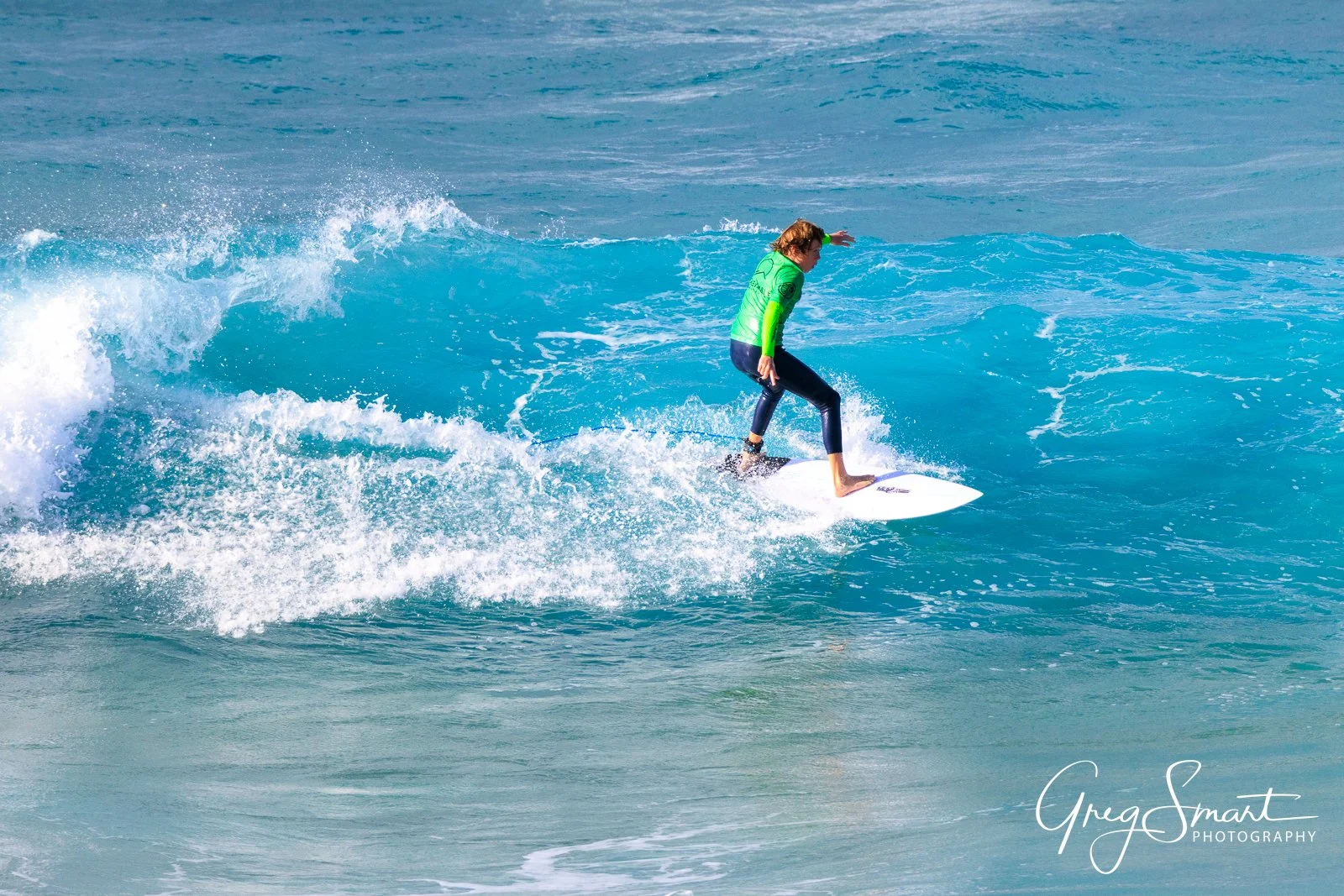 A person surfing on a blue wave in the ocean, wearing a green long-sleeve shirt and dark pants.