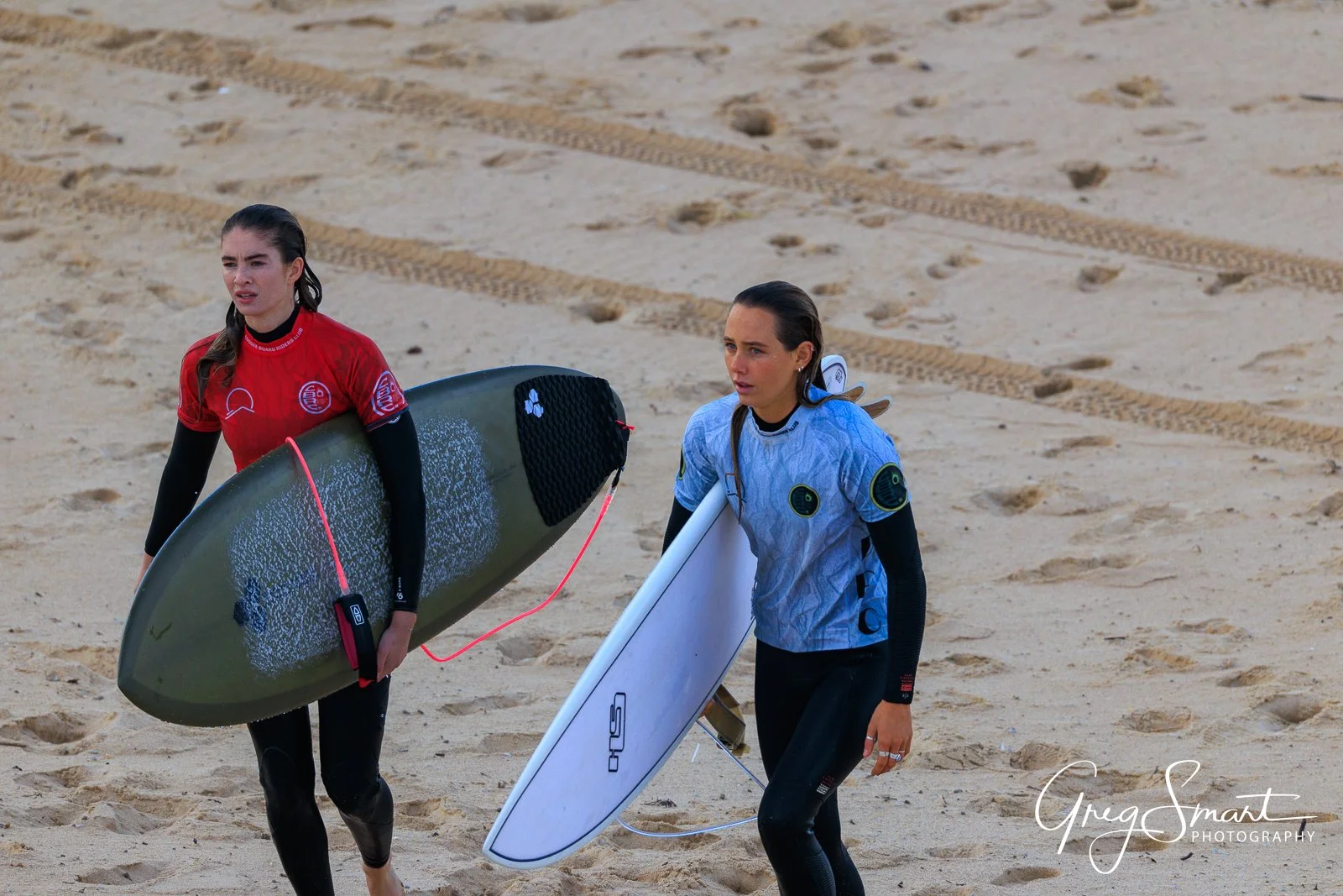 Two female surfers walking on sandy beach, each carrying a surfboard, with tire tracks and foot prints in the sand.