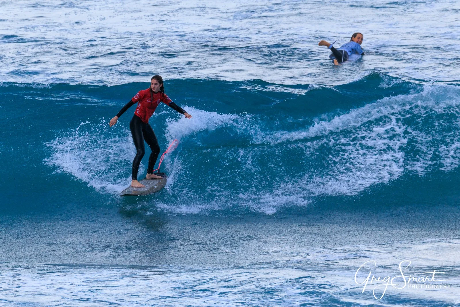 A woman surfboarding on a wave with a man swimming behind her in the ocean.
