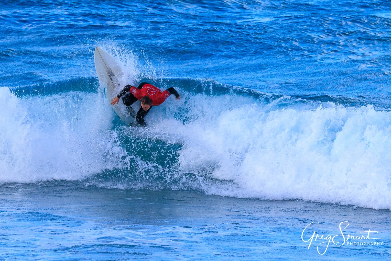 A surfer wearing a black wetsuit and red jersey riding a wave on a surfboard in the ocean.