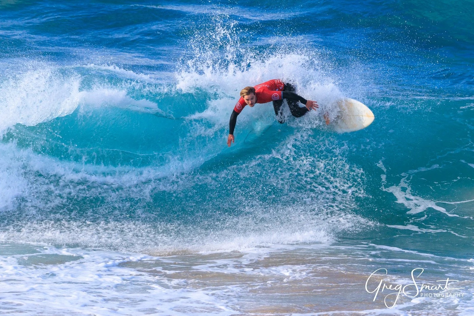 A surfer in a red wetsuit riding a blue wave at the beach.
